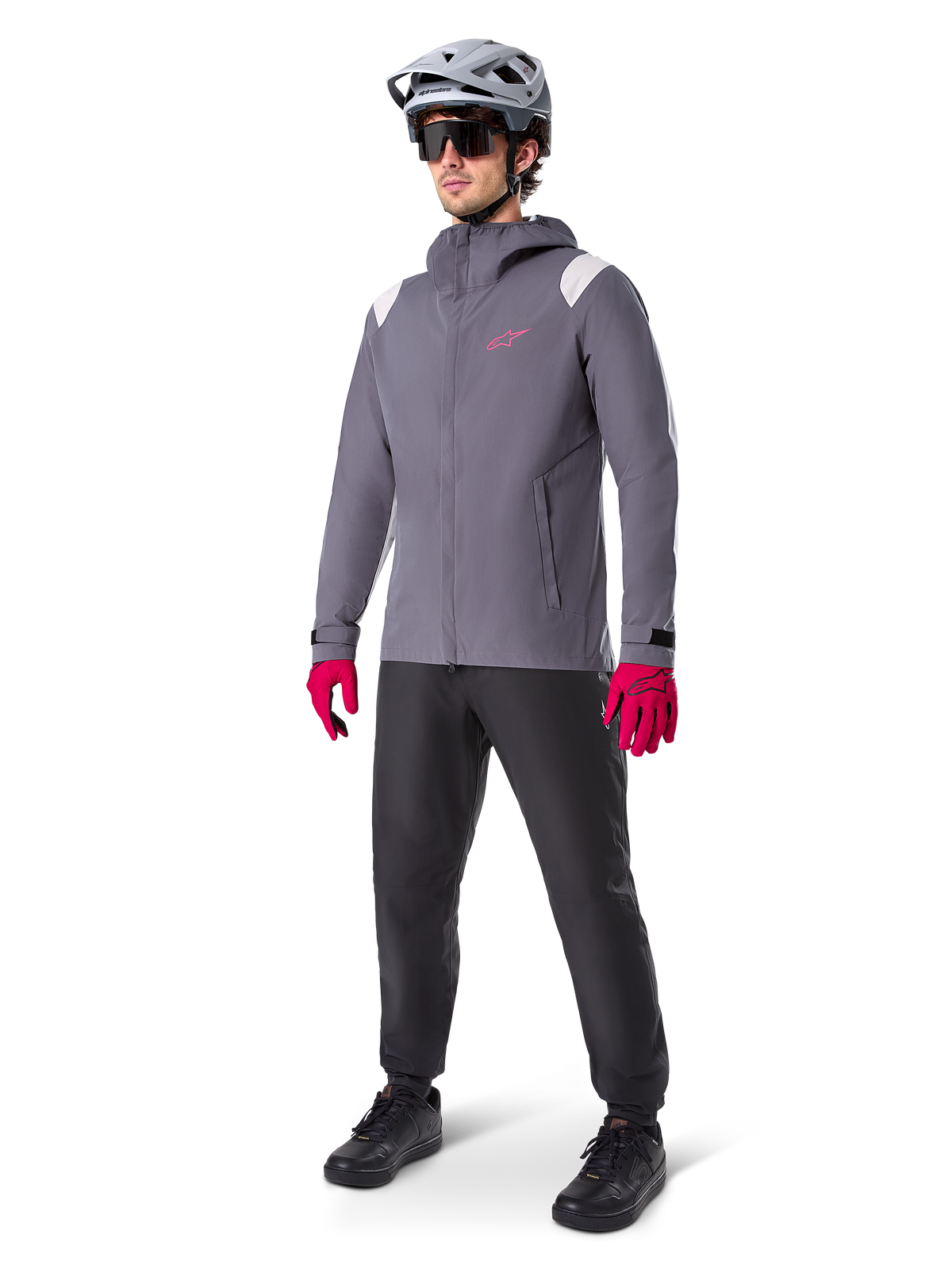 Modello che indossa la giacca antipioggia Alpinestars A-Dura in Dark Titanium, giacca grigia con cappuccio, logo rosa sul petto e inserti grigio chiaro sulle spalle, abbinata a un casco da MTB grigio, occhiali da sole scuri, guanti rosa vivace e pantaloni neri per ciclismo fuoristrada.
