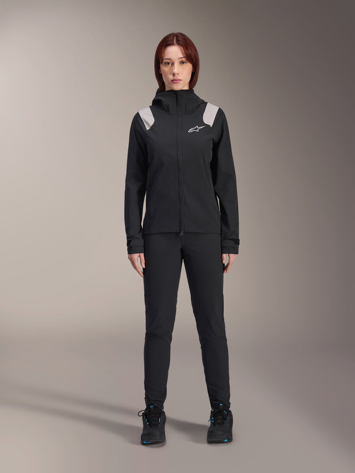 Alpinestars Stella A-Dura Rain Jacket, giacca antipioggia MTB da donna, nera con accenti grigio chiaro sulle spalle e logo bianco, indossata con pantaloni e scarpe neri abbinati, dotata di cappuccio e polsini regolabili, progettata per il trail riding in mountain bike