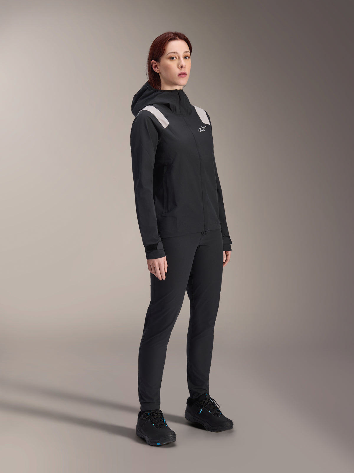 Donna che indossa la giacca antipioggia Alpinestars Stella A-Dura in nero, guscio MTB impermeabile a 2,5 strati con dettagli grigio chiaro sulle spalle, pantaloni neri e scarpe da ciclismo abbinati, ritratto in studio per abbigliamento tecnico da ciclismo