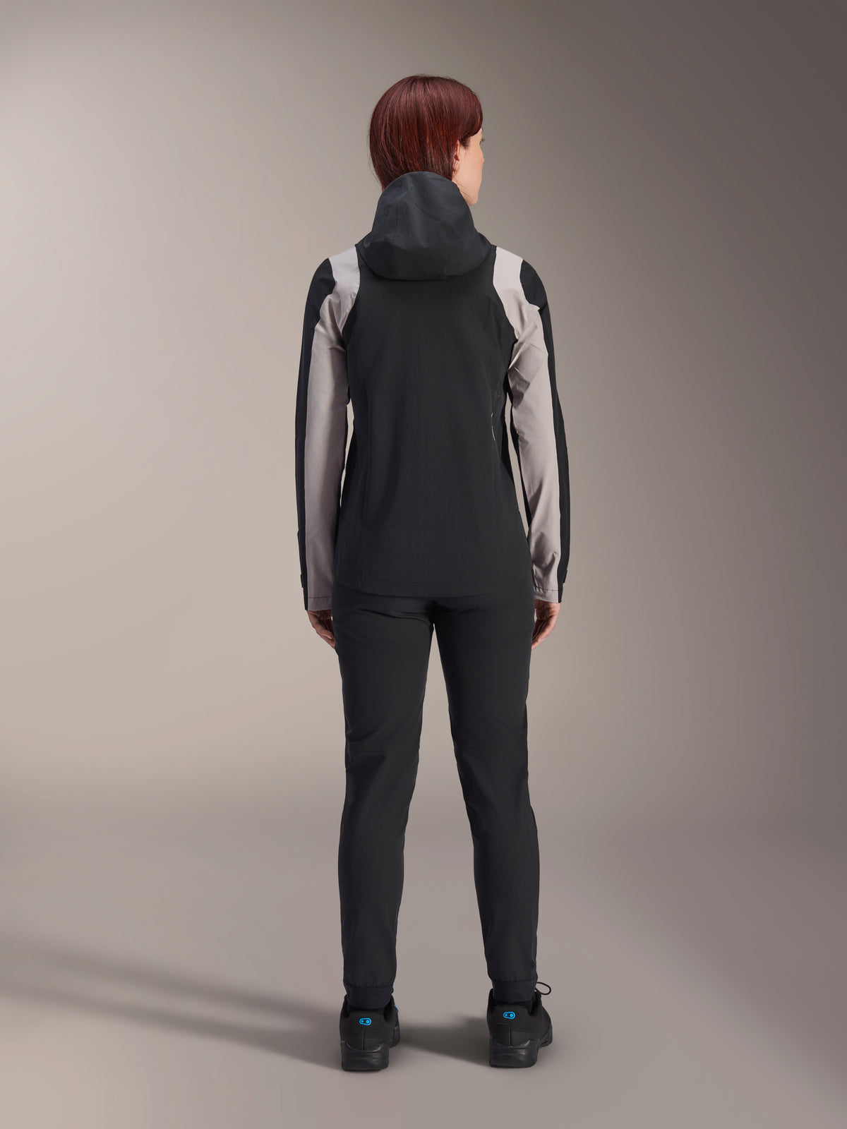 Alpinestars Stella A-Dura Rain Jacket, ciclista vista da dietro, nera con inserti grigio chiaro su maniche e spalle, cappuccio sollevato, indossata con pantaloni neri e scarpe da ciclismo, abbigliamento tecnico antipioggia per mountain bike