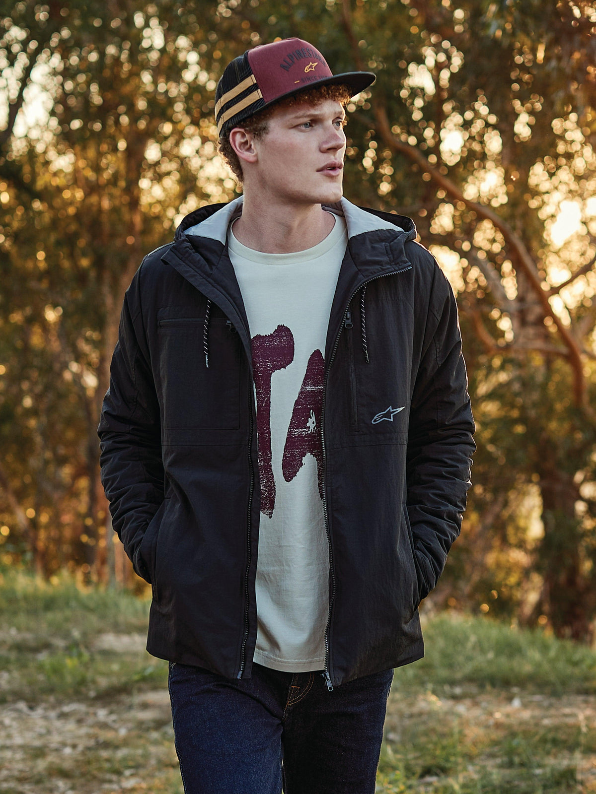 Cappello Trucker Alpinestars Backline in color mattone, nero e senape, indossato da un uomo con giacca nera con cappuccio e t-shirt grafica, streetwear casual ispirato al motorsport in un ambiente all'aperto alla luce del tramonto.