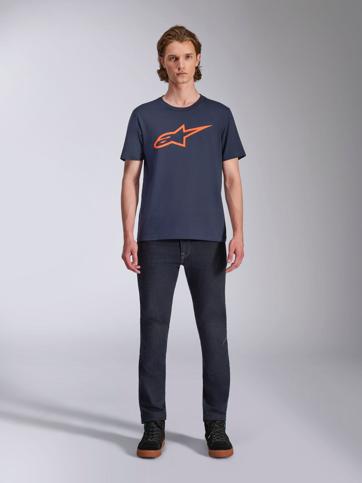 Alpinestars Ageless 2.0 CSF Tee, maglietta a maniche corte, blu navy con logo Alpinestars arancione, indossata da un modello con jeans scuri e sneakers, streetwear casual da motorsport
