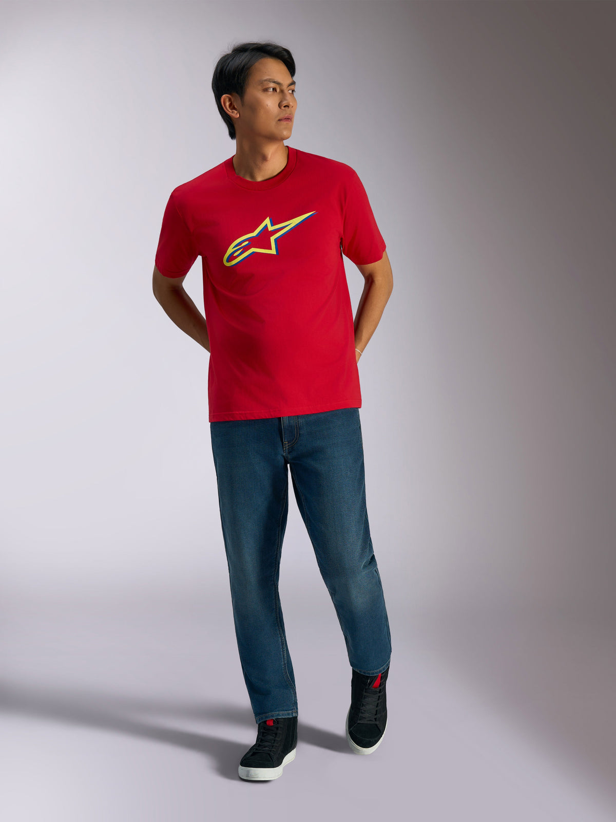 Alpinestars Ageless Shadow CSF Tee, t-shirt a maniche corte rossa, grafica con logo in ombra giallo e blu, indossata con jeans blu e sneakers nere, abbigliamento casual motorsport lifestyle