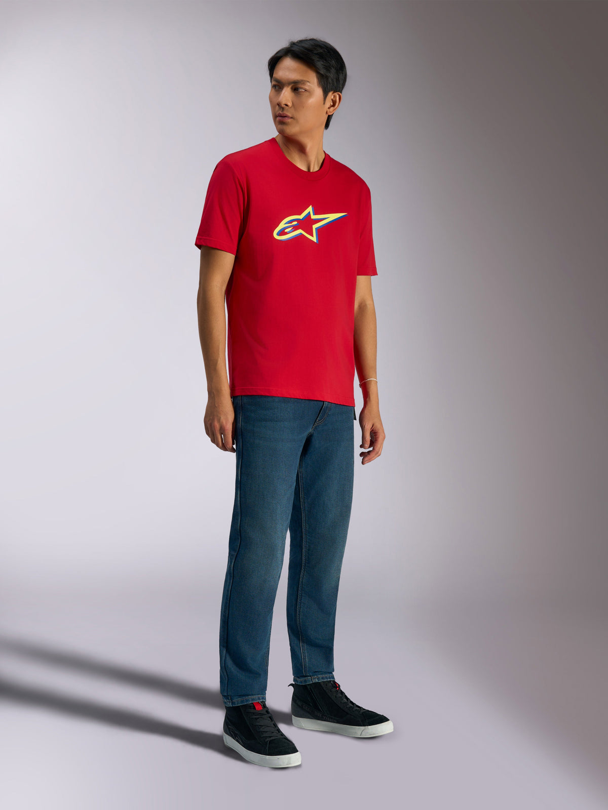 Alpinestars Ageless Shadow CSF Tee, t-shirt a maniche corte, rossa, con logo Ageless shadow e accenti gialli e blu, indossata da un modello con jeans in denim e sneakers nere, abbigliamento casual motorsport streetwear