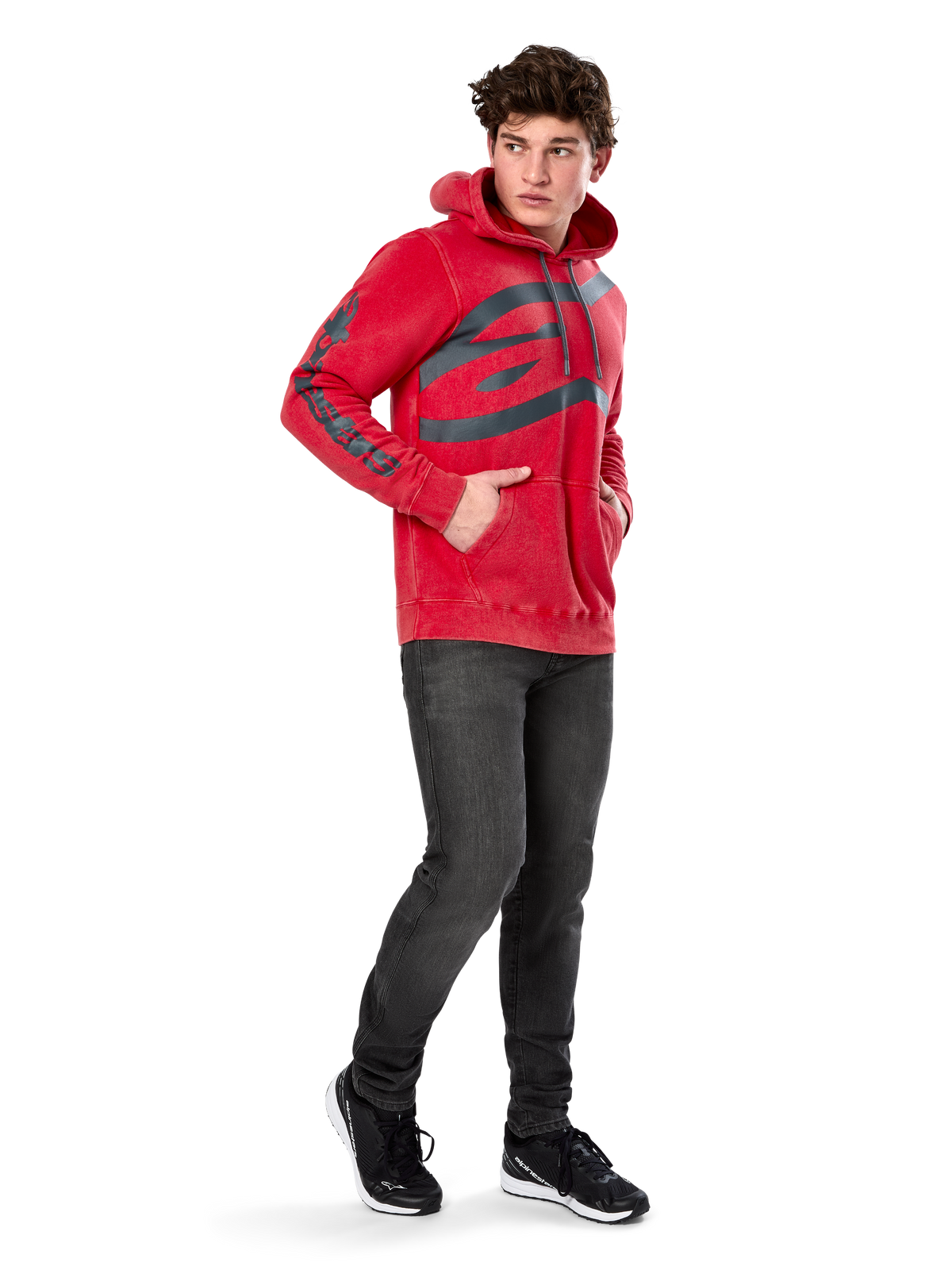 Alpinestars Unbound Hoodie, felpa in pile rossa tinta in capo, logo scuro extra large sul petto e scritta sulle maniche, tasca a marsupio, cappuccio con cordino, indossata da un modello con jeans grigio scuro e sneakers nere, streetwear casual ispirato al motorsport