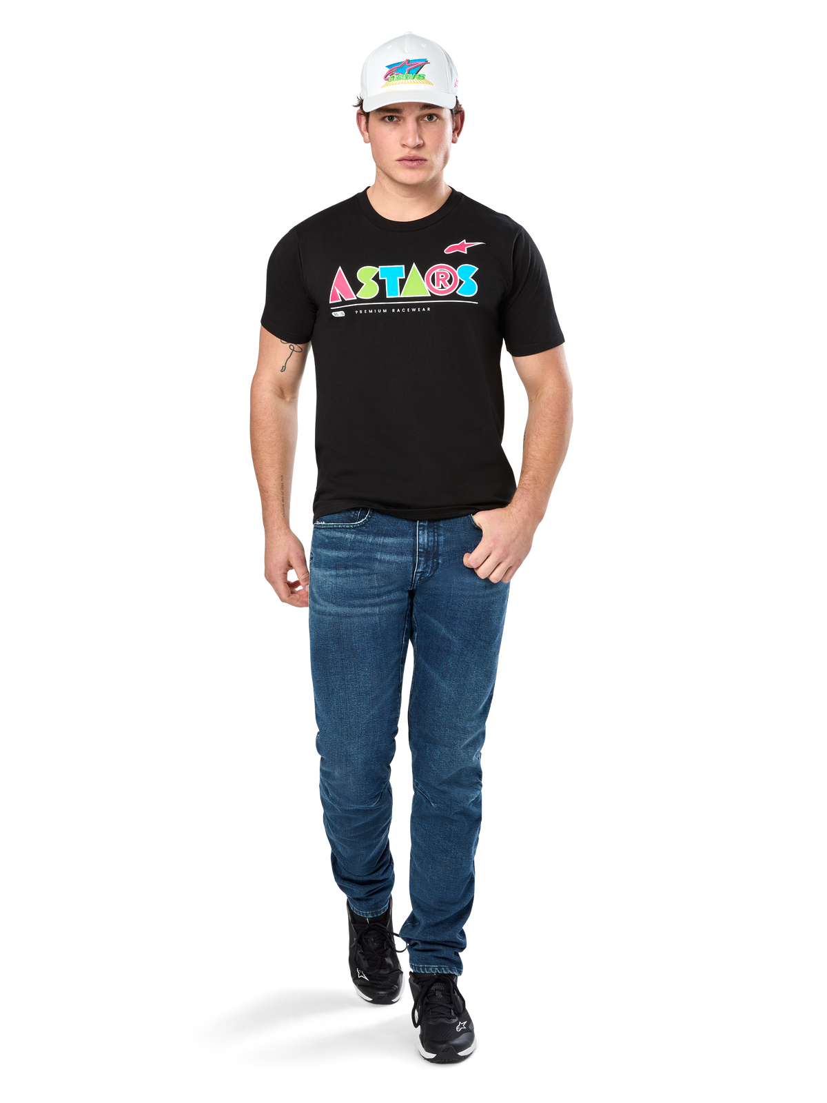 Modello maschile che indossa la maglietta Alpinestars Cereal CSF, T-shirt nera a maniche corte con logo ASTARS geometrico colorato, abbinata a jeans blu e cappellino bianco, streetwear casual ispirato al motorsport