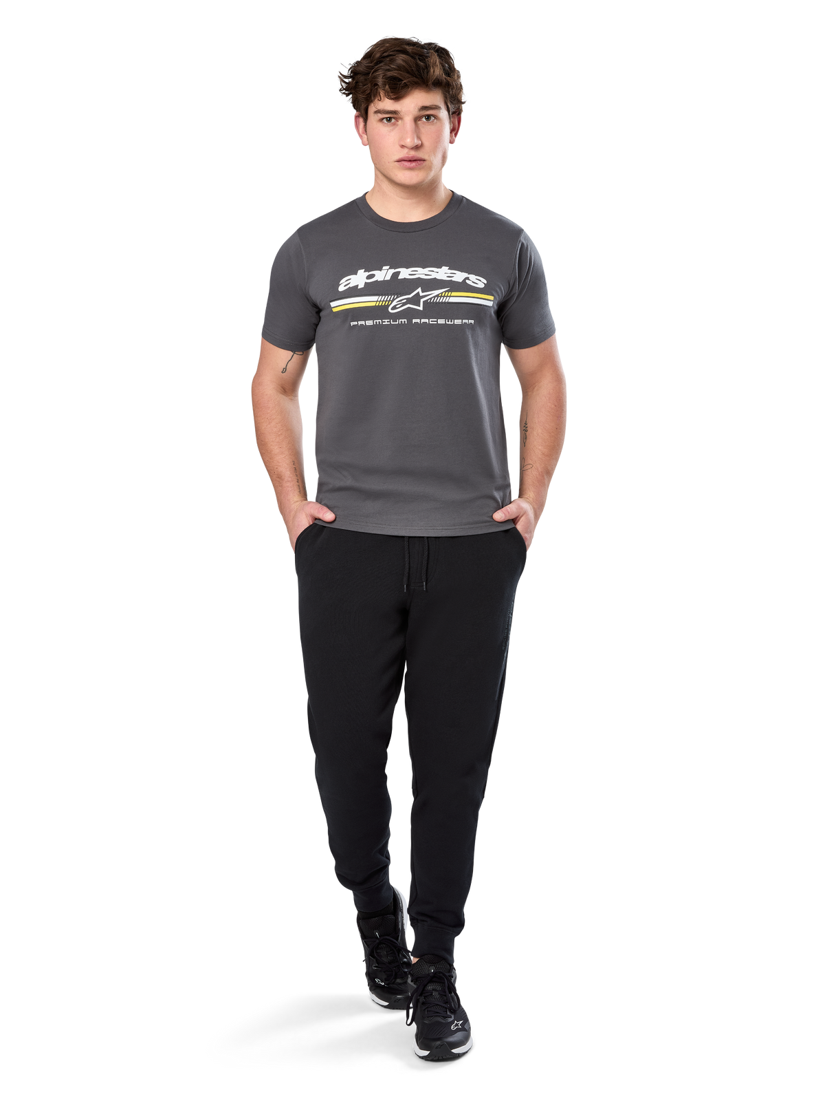 Uomo che indossa la Alpinestars Prevail Csf Tee, t-shirt a maniche corte, grigio antracite, con logo Alpinestars bianco e striscia tricolore con accenti gialli, abbinata a pantaloni sportivi neri e sneakers, abbigliamento casual motorsport streetwear