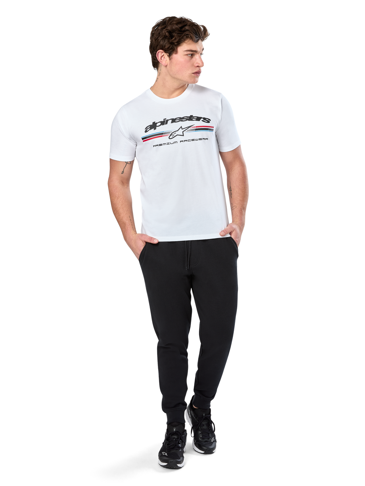 Alpinestars T-shirt Prevail CSF, t-shirt a maniche corte, bianca, con logo a strisce tricolore stile paddock anteriore e marchio Alpinestars, indossata da un modello con pantaloni sportivi neri e sneaker nere, streetwear casual da motorsport