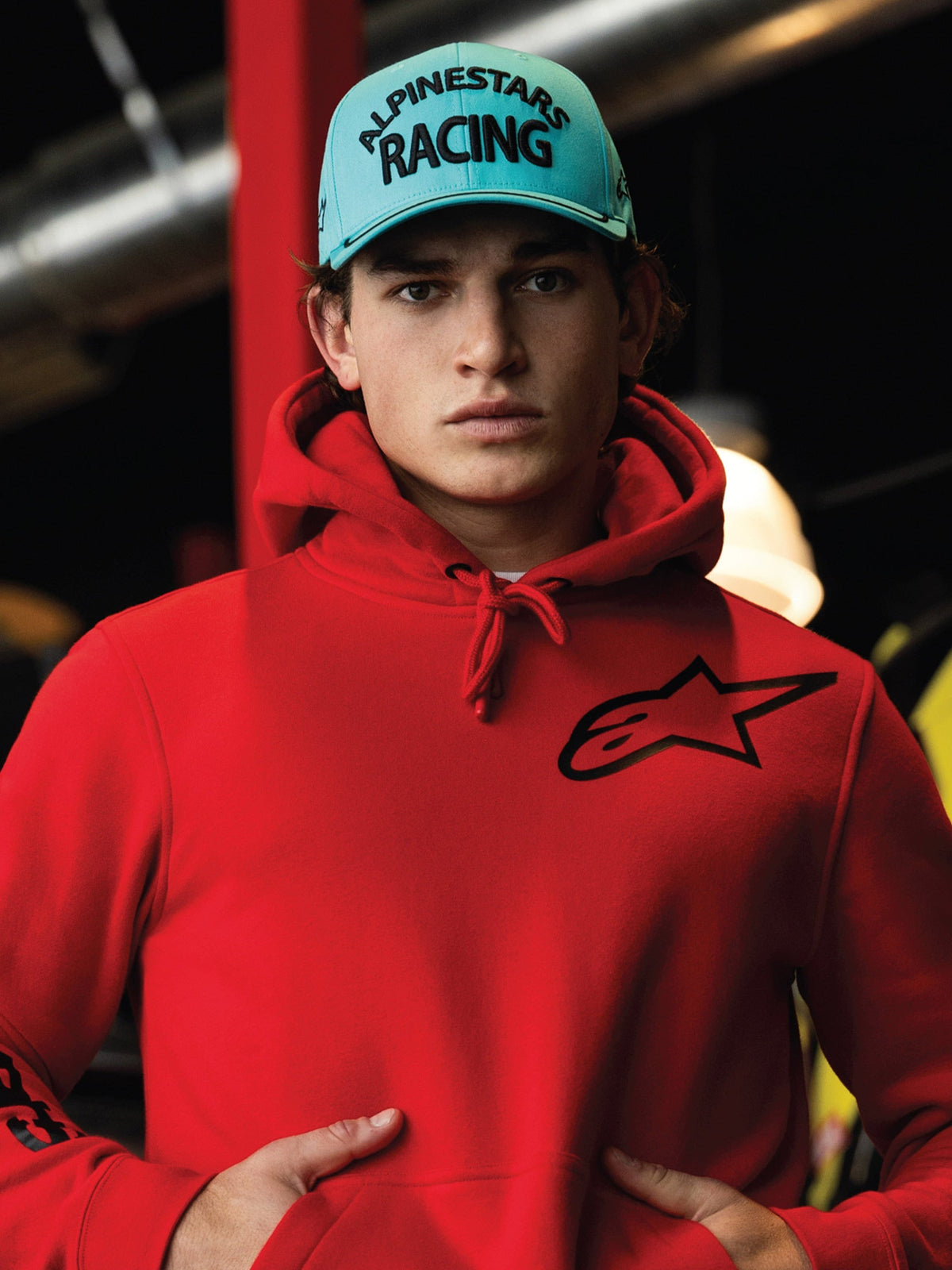 Cappello Alpinestars Luminary, snapback blu acqua con visiera curva, ricamo 3D nero con scritta 'ALPINESTARS RACING', indossato con una felpa rossa Alpinestars, streetwear casual in stile motorsport