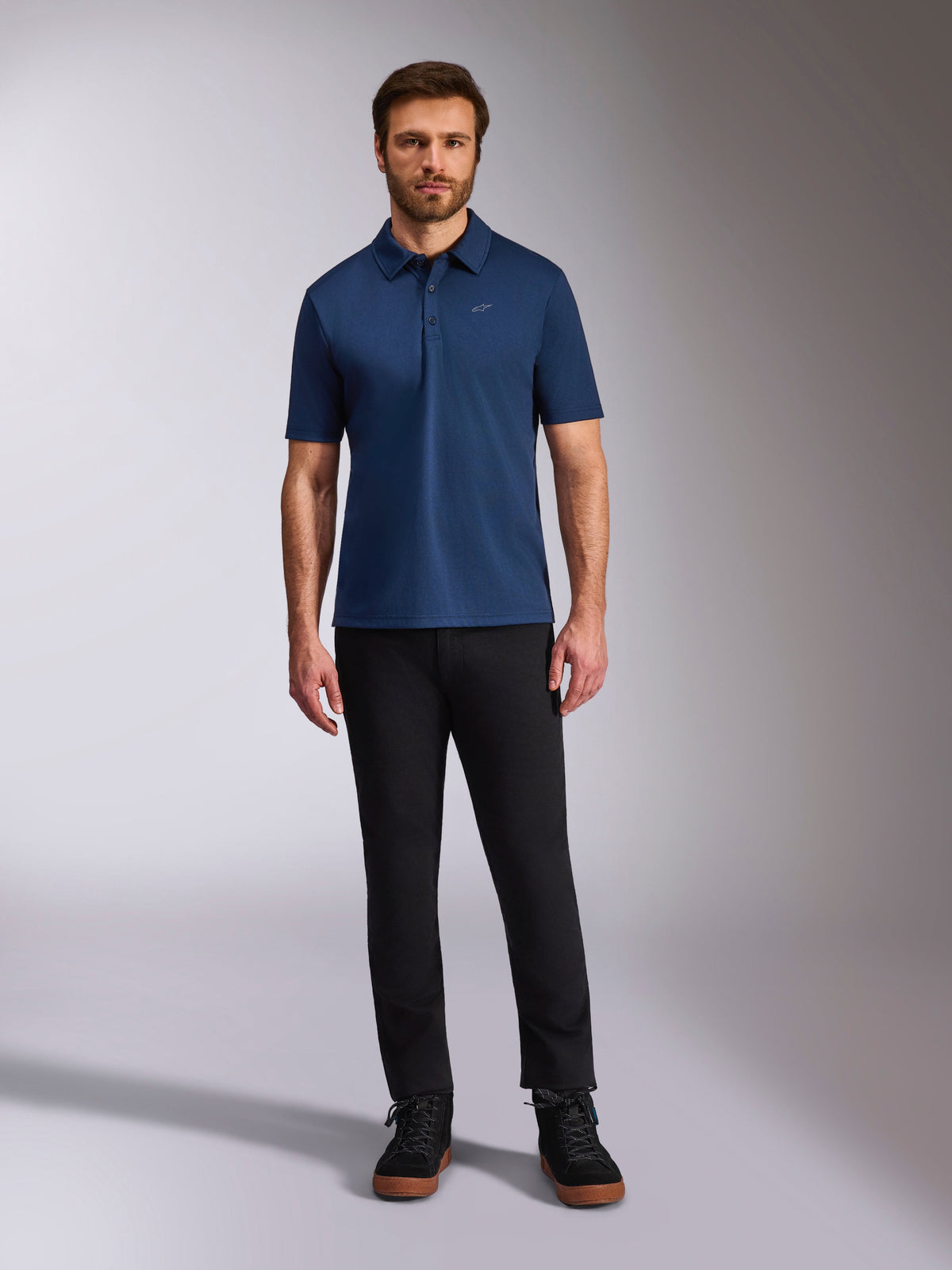 Alpinestars Impetus Sport Polo, polo a maniche corte, blu navy, piccolo logo Astars argento sul petto, abbottonatura a tre bottoni, indossata da un modello con pantaloni neri e stivali, abbigliamento sportivo casual ispirato al motorsport