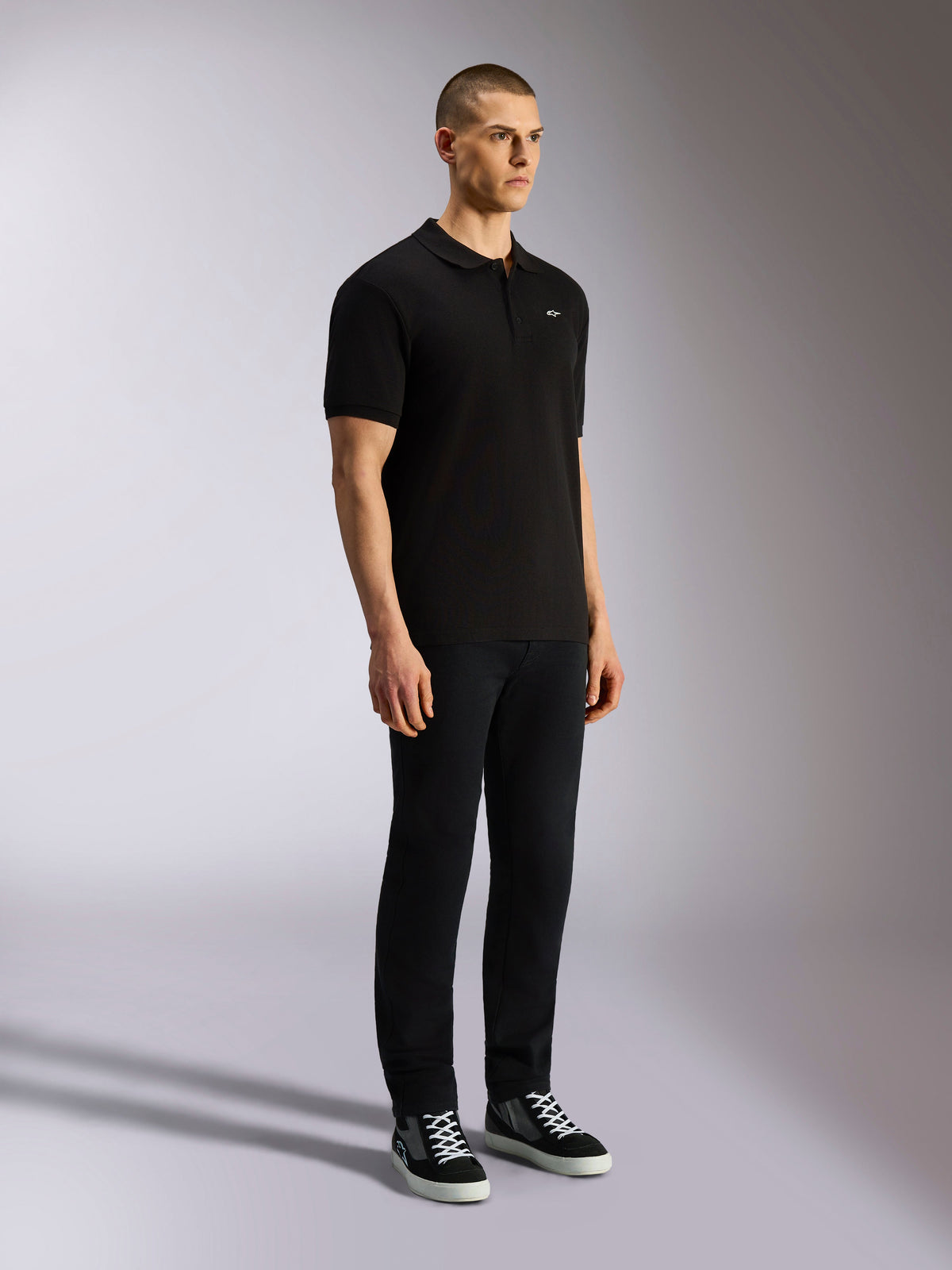 Alpinestars Endeavor Classic Polo, polo nera a maniche corte, tessuto piqué con abbottonatura a tre bottoni, piccolo logo Astars bianco sul petto a sinistra, indossata con pantaloni neri e sneakers, streetwear casual motorsport