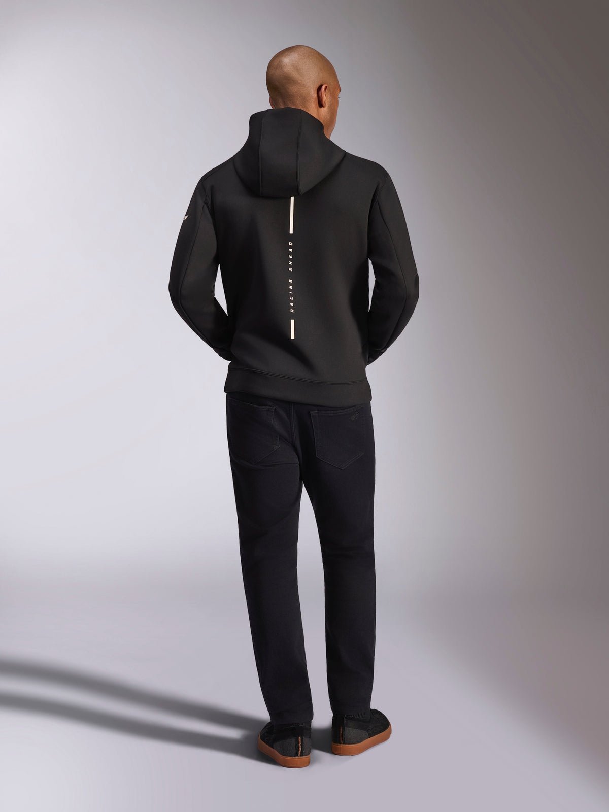 Alpinestars Insight Hoodie, nero, vista posteriore su modello, caratterizzata da un logo a striscia bianca verticale sul retro con scritta "Racing Ahead", felpa sportiva casual in pile double face tri-blend, abbinata a pantaloni neri.