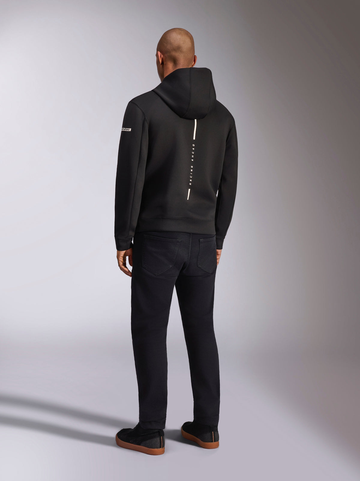 Alpinestars Insight Hoodie, felpa sportiva nera, vista posteriore con striscia bianca verticale e branding 'RACING AHEAD', logo sulla manica, abbinata a pantaloni neri e sneakers, design casual ispirato al motorsport