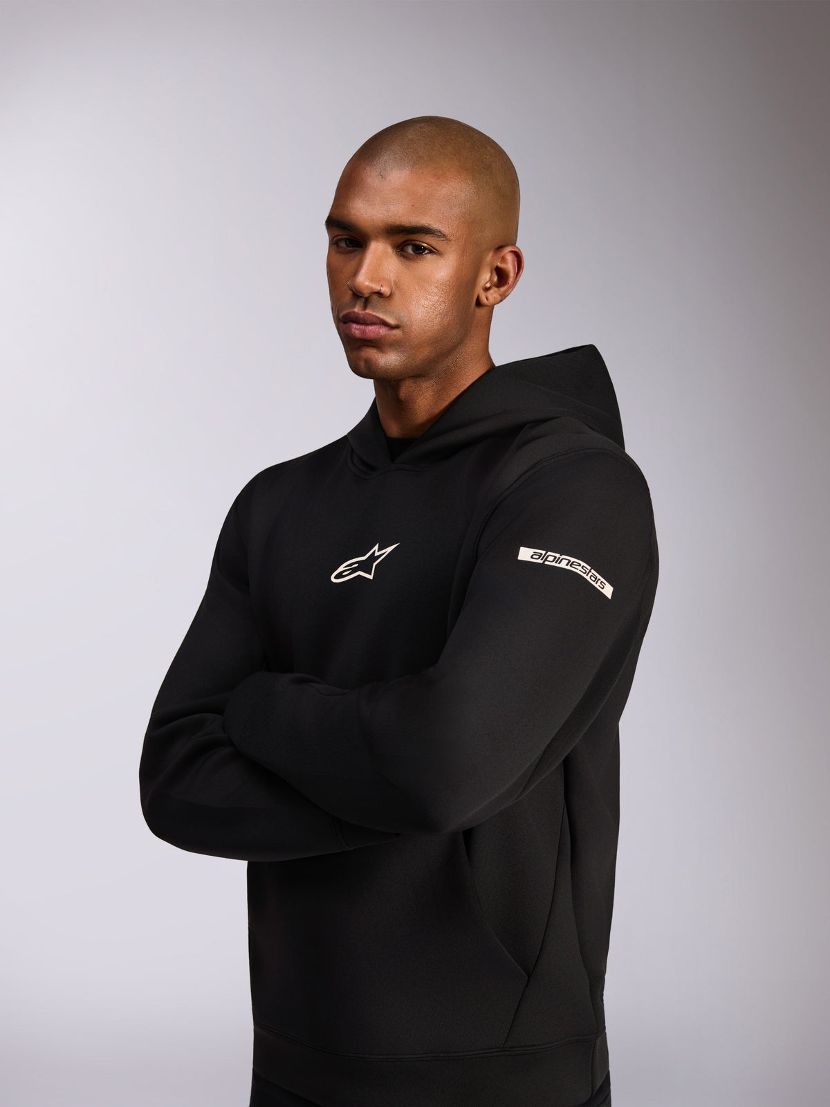 Alpinestars Insight Hoodie, felpa sportiva casual, nera, logo Astars bianco sul petto e logo Alpinestars Word sulla manica, modello che indossa la felpa con le braccia incrociate, pregiata costruzione in pile double face
