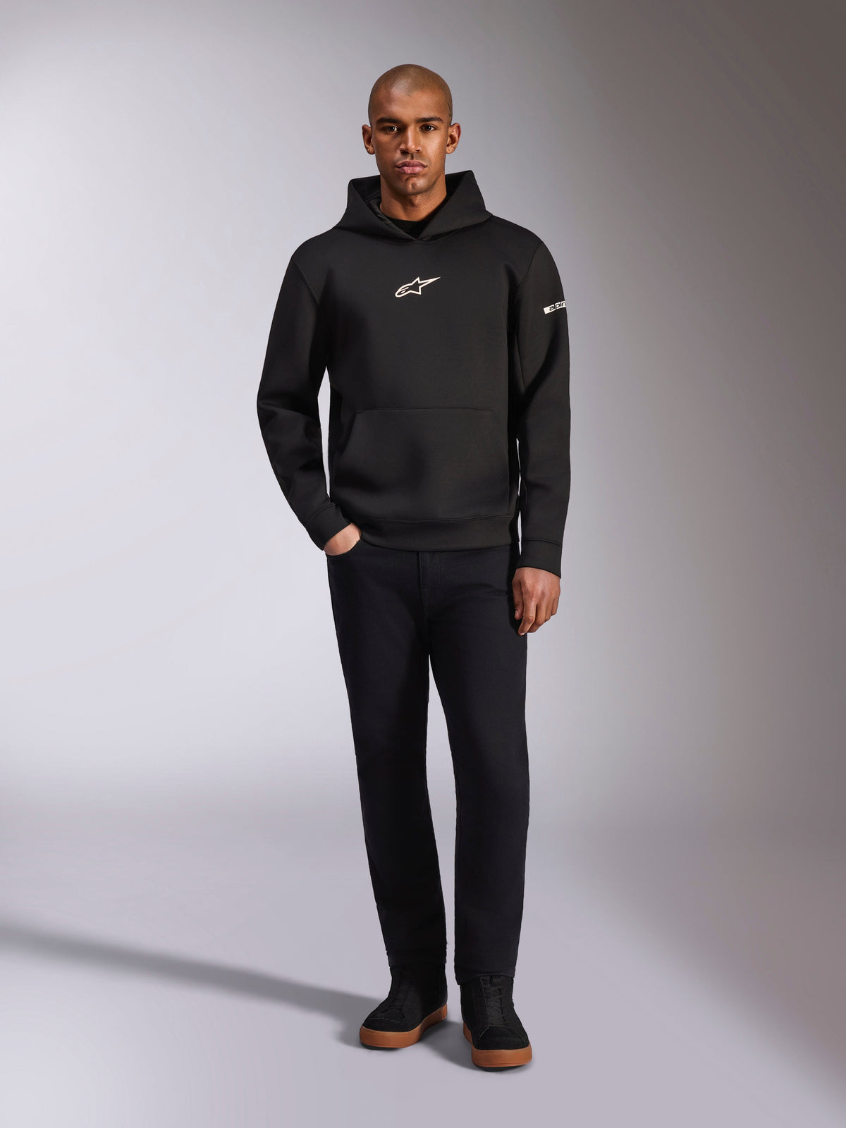 Alpinestars Insight Hoodie, felpa nera con cappuccio e logo Astars bianco sul petto e sulla manica, indossata da un modello con pantaloni neri e sneakers nere, streetwear casual ispirato al motorsport