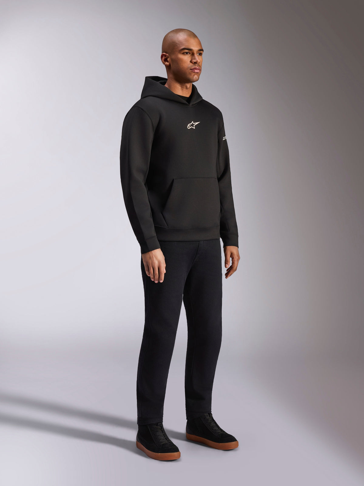 Alpinestars Insight Hoodie, nero, felpa casual pullover in pile con logo Astars bianco sul petto, tasca a marsupio, indossata con pantaloni neri e sneakers, scatto in studio a figura intera, streetwear ispirato al motorsport