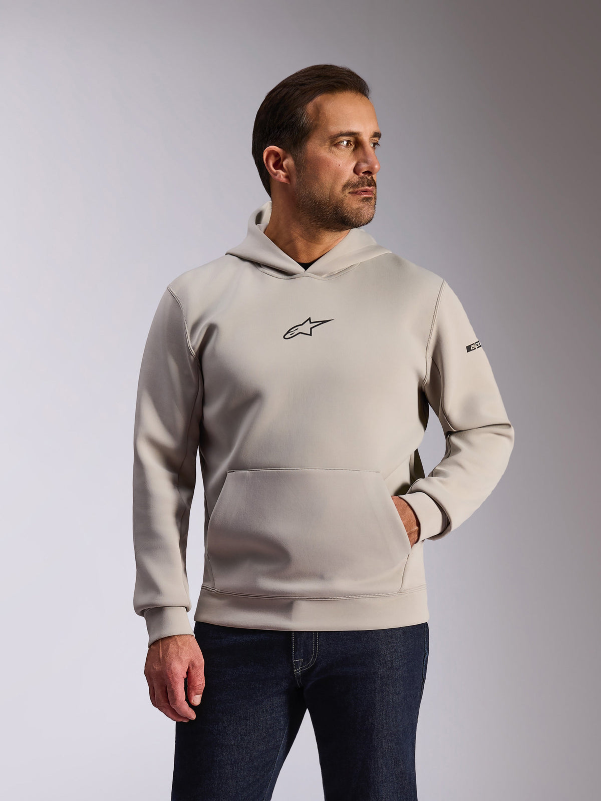 Felpa con cappuccio Alpinestars Insight, abbigliamento sportivo casual, grigio cemento, design a pullover con tasca a marsupio e piccolo logo Astars nero sul petto, maniche lunghe con logo Alpinestars sulla manica sinistra, indossata da un modello con jeans in denim scuro, abbigliamento lifestyle motorsport