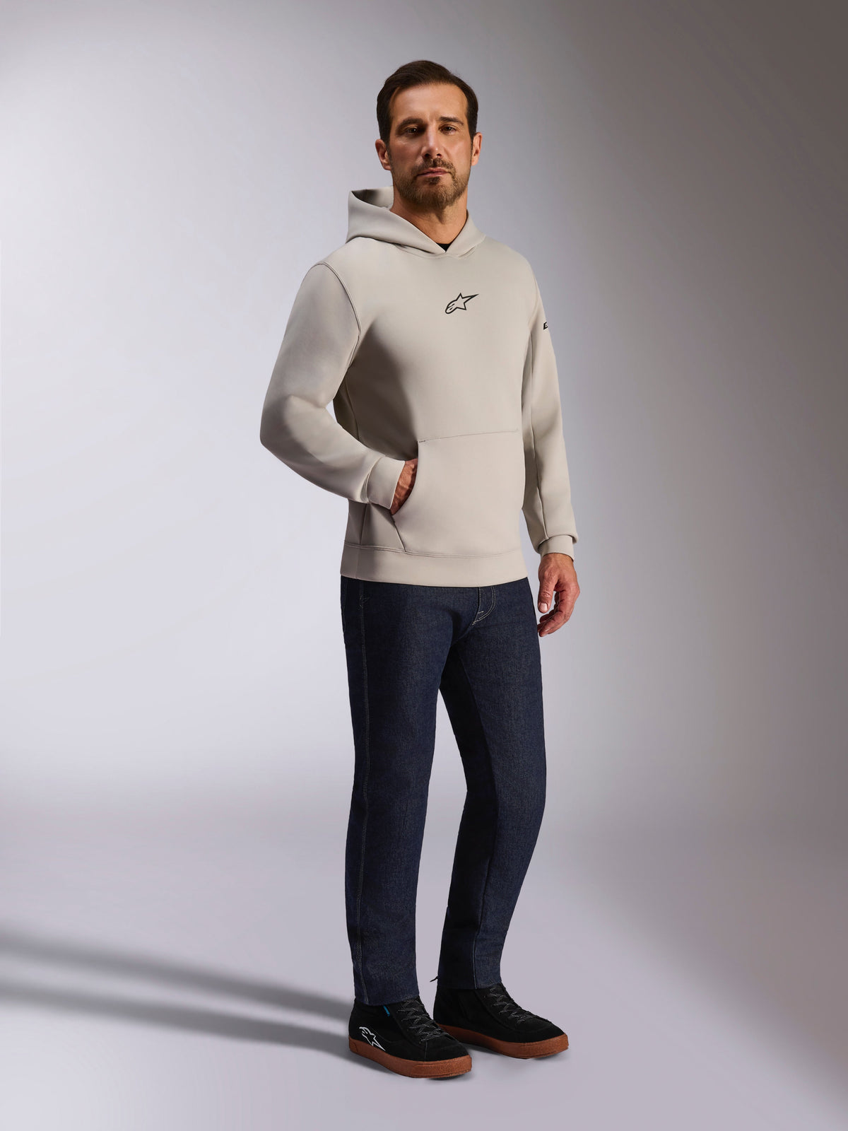 Felpa con cappuccio Alpinestars Insight, streetwear casual, grigio cemento con logo Astars nero sul petto, modello pullover con tasca a marsupio, indossata con jeans blu scuro e sneakers nere, stile moderno ispirato al motorsport