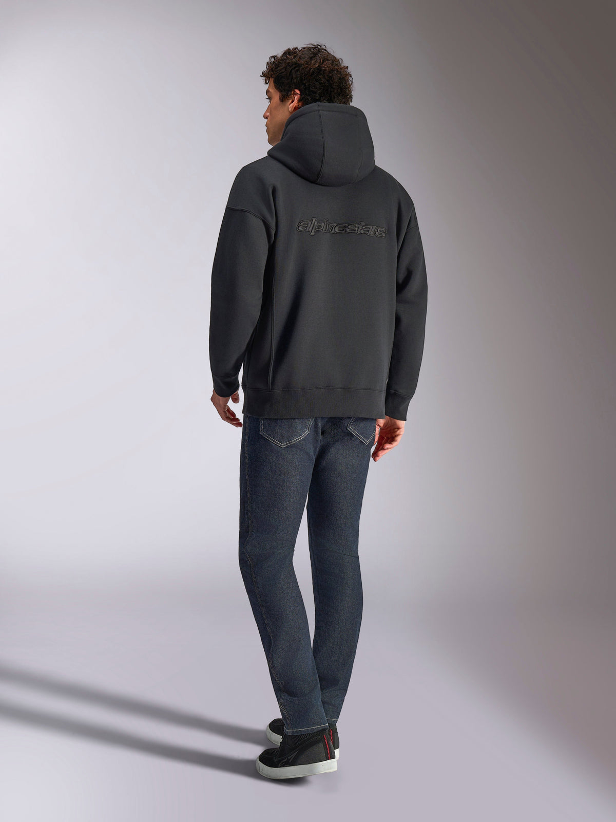 Felpa con cappuccio Alpinestars Emergent Oversized, nero, vista posteriore con logo Alpinestars in ricamo 3D sulle spalle, vestibilità oversize, abbinata a jeans in denim blu scuro e sneaker nere, streetwear casual ispirato al motorsport
