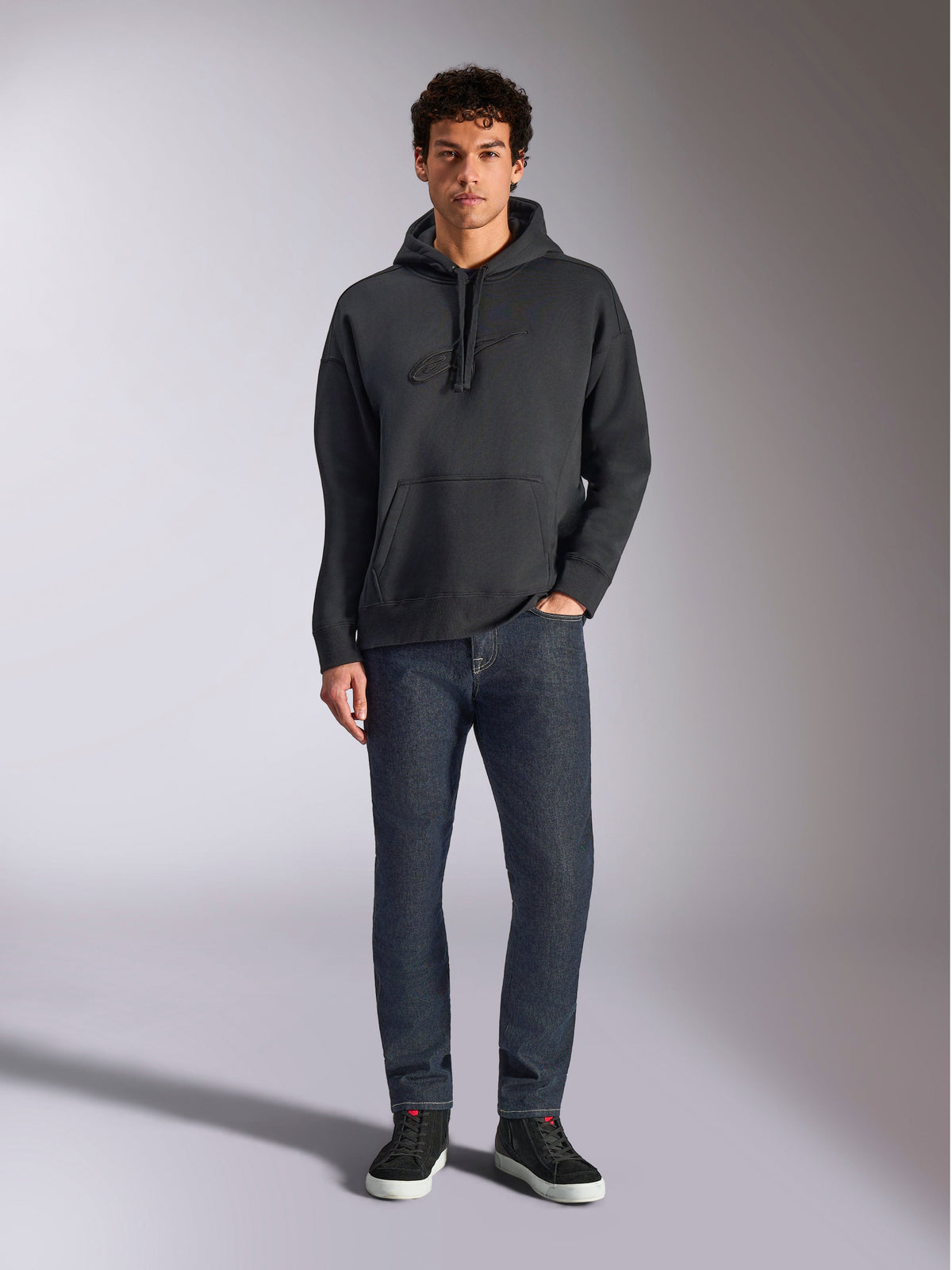 Alpinestars Emergent Oversized Hoodie, abbigliamento lifestyle streetwear casual, nero con logo ricamato in 3D tono su tono, cappuccio con cordino, tasca a marsupio, vestibilità oversize, indossata da un modello con jeans blu scuro e sneakers nere