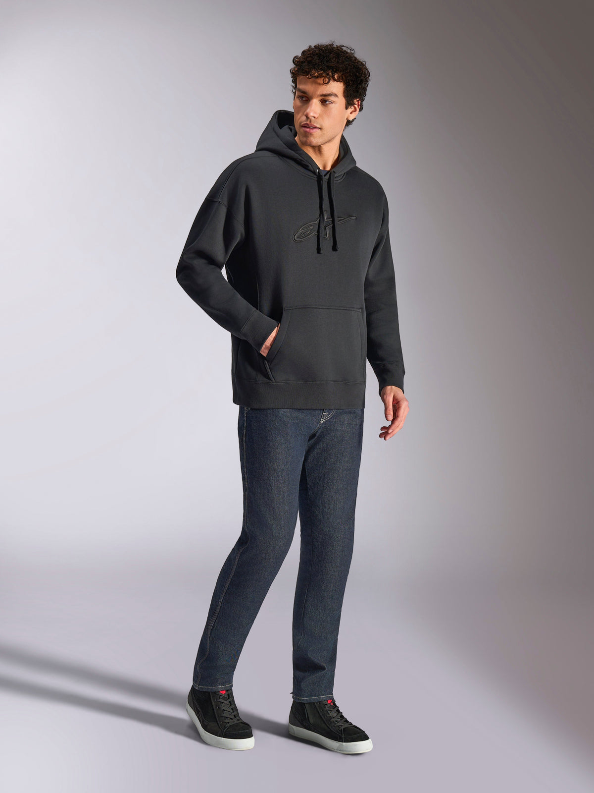 Alpinestars Emergent Oversized Hoodie, felpa casual streetwear nera, logo Astars ricamato in 3D tono su tono sul petto, tasca a marsupio, vestibilità oversize, indossata da un modello con jeans in denim blu scuro e sneakers nere