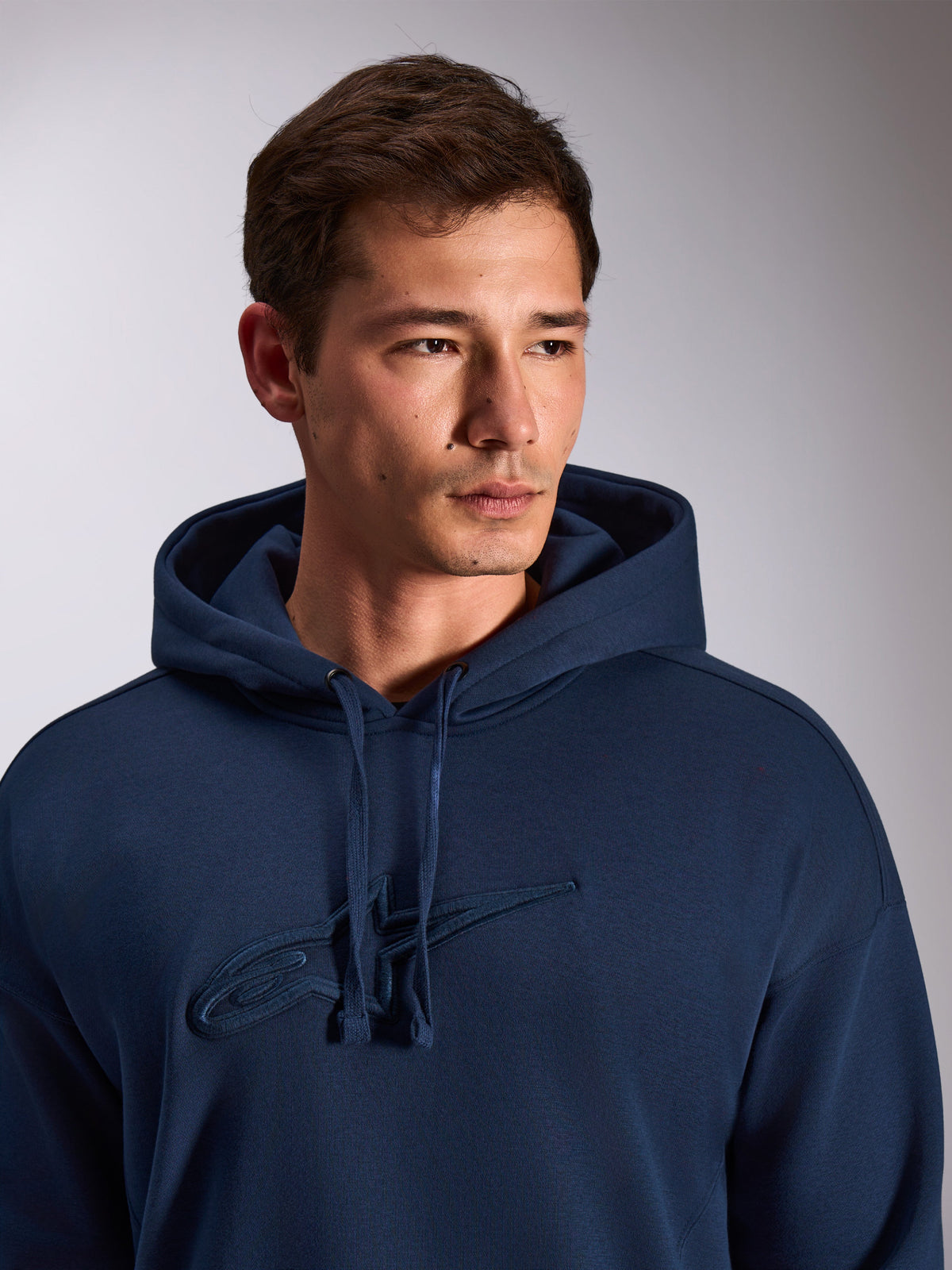 Felpa con cappuccio Alpinestars Emergent Oversized, blu navy, primo piano di un modello che indossa una felpa in pile, con logo Astars ricamato in 3D tono su tono sul petto, cappuccio con cordino regolabile, streetwear casual ispirato al motorsport