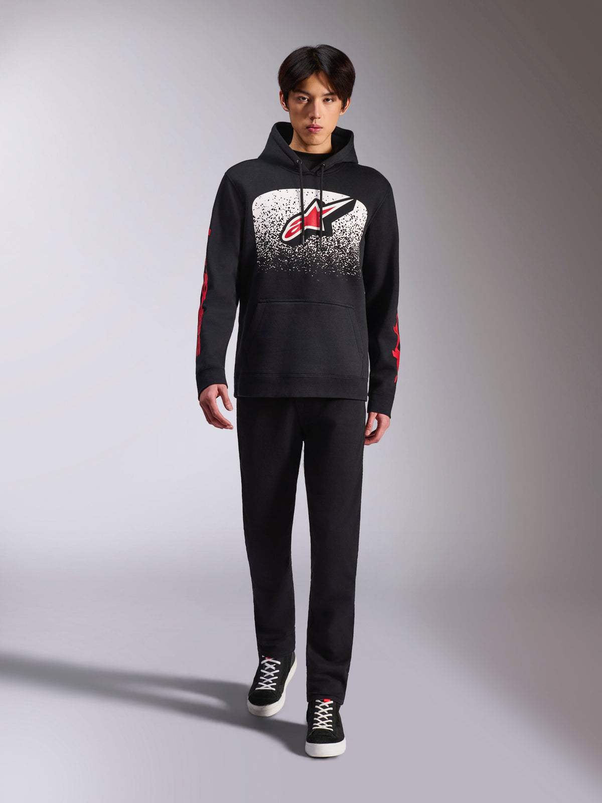 Alpinestars Obsessed Hoodie, felpa nera con cappuccio, grande logo Astars bianco e rosso con gradazione MX, scritte rosse sulle maniche, indossata da un modello con outfit casual total black, abbigliamento lifestyle ispirato al motorsport