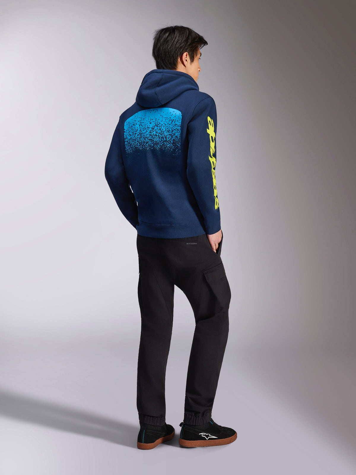 Alpinestars Obsessed Hoodie, felpa casual blu navy, vista posteriore con grafica MX sfumata azzurra e logo Alpinestars giallo sulla manica, indossata con pantaloni neri e sneakers