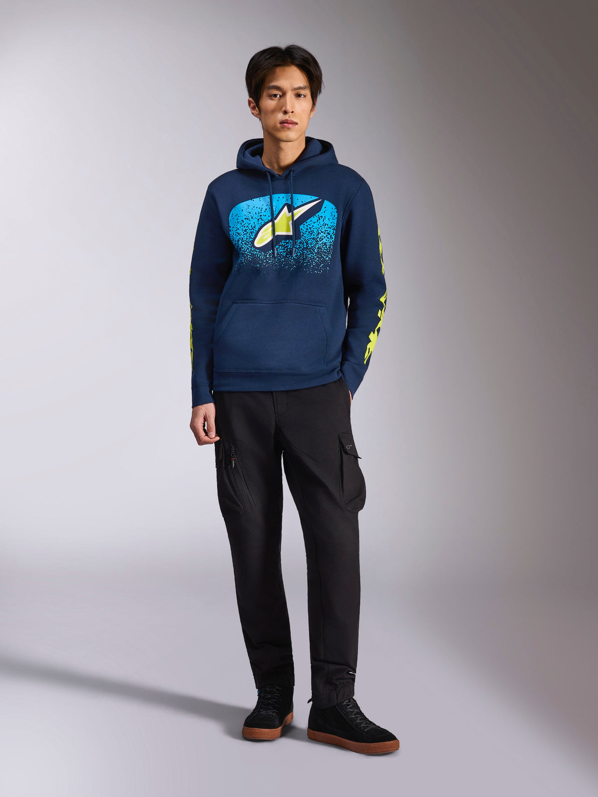 Felpa Alpinestars Obsessed Hoodie, felpa pullover blu navy, logo Astars frontale con sfumatura MX blu e stella giallo lime, loghi Alpinestars giallo lime sulle maniche, indossata da un modello con pantaloni cargo neri e sneakers nere, streetwear casual ispirato al motorsport