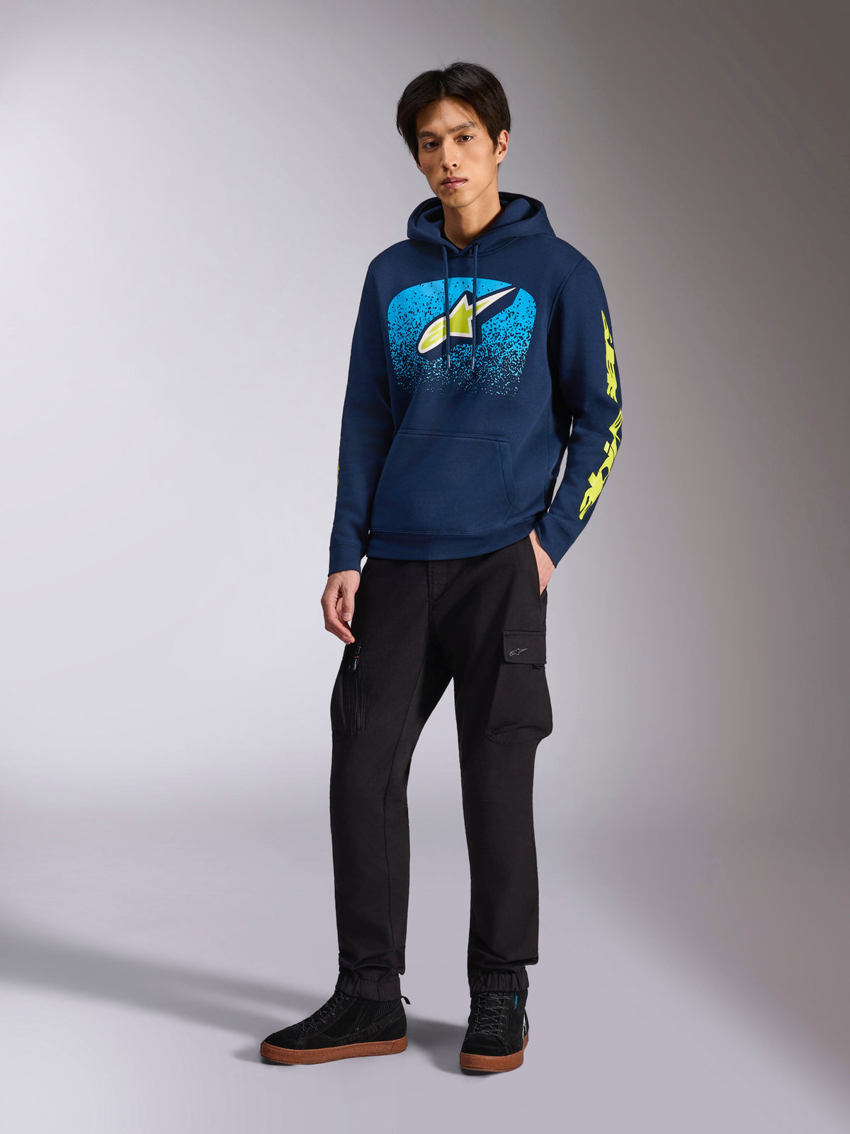 Alpinestars Obsessed Hoodie Navy, felpa casual con grafica frontale azzurra e giallo lime, branding Alpinestars giallo lime sulle maniche, indossata da un modello con pantaloni cargo neri e sneakers nere, streetwear ispirato al motorsport