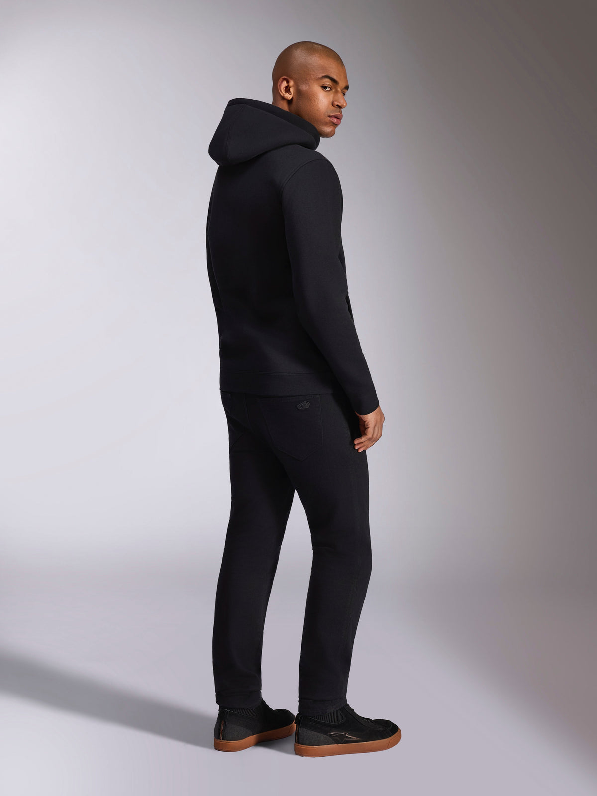 Alpinestars Felpa con cappuccio Ellipsoid, streetwear casual nero, vista posteriore su modello, felpa pullover con cappuccio, polsini e orlo a coste, indossata con pantaloni neri e sneakers, design moderno ispirato al motorsport