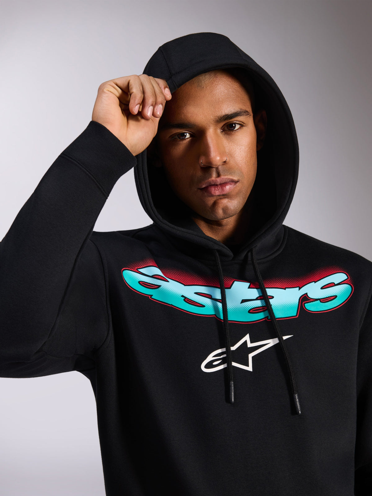 Alpinestars Ellipsoid Hoodie, felpa in pile nera a pullover, con scritta Astars stilizzata e sfumata verde acqua e rosso e logo bianco sul petto, indossata da un modello con cappuccio alzato, streetwear casual ispirato al motorsport