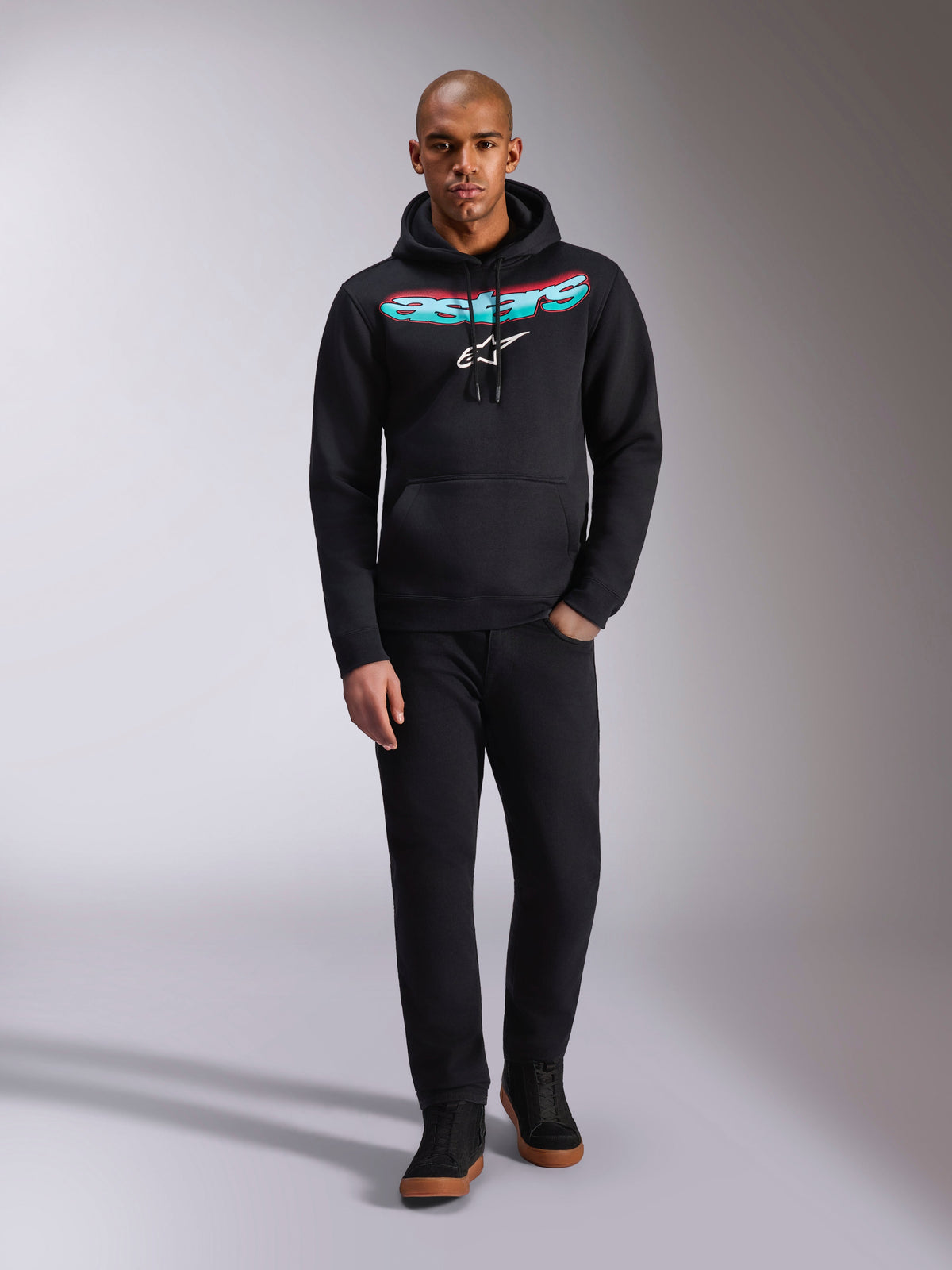 Felpa con cappuccio Alpinestars Ellipsoid, nera, felpa in pile modello pullover con scritta Astars curva con sfumatura di colore rosso e verde acqua e logo a stella bianco, indossata da un modello con pantaloni neri e sneakers, streetwear casual motorsport