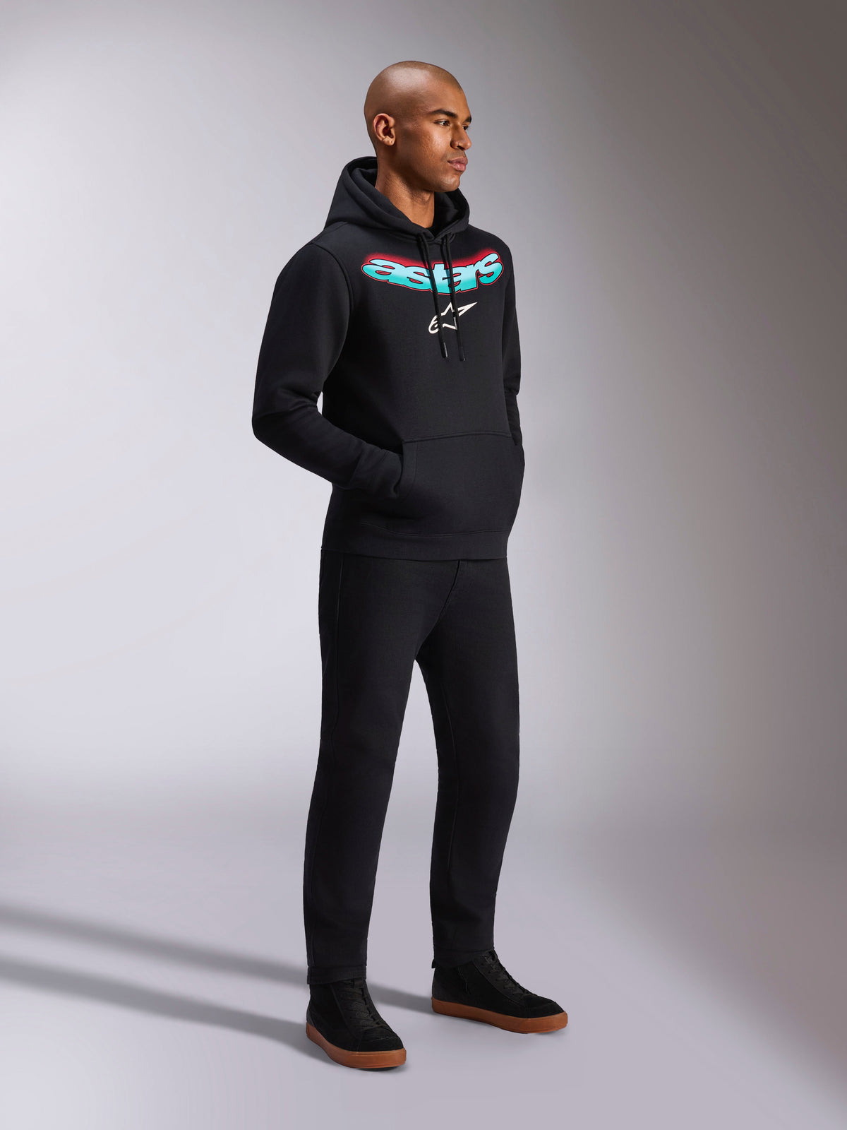Felpa con cappuccio Alpinestars Ellipsoid, pullover in pile nero, con scritta Astars curva in gradiente verde acqua e rosso e logo a stella bianco, indossata da un modello con pantaloni neri, streetwear casual ispirato al motorsport