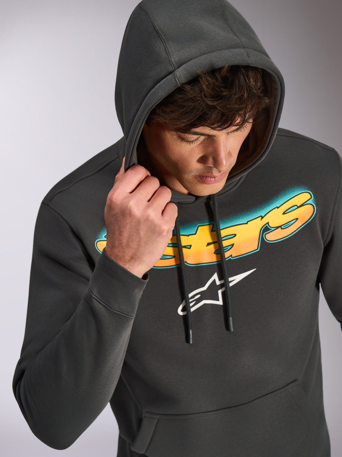 Felpa Alpinestars Ellipsoid, grigio canna di fucile, felpa pullover casual con scritta Astars sfumata dal ciano all'arancione e logo bianco, indossata dal modello con il cappuccio alzato, streetwear ispirato al motorsport