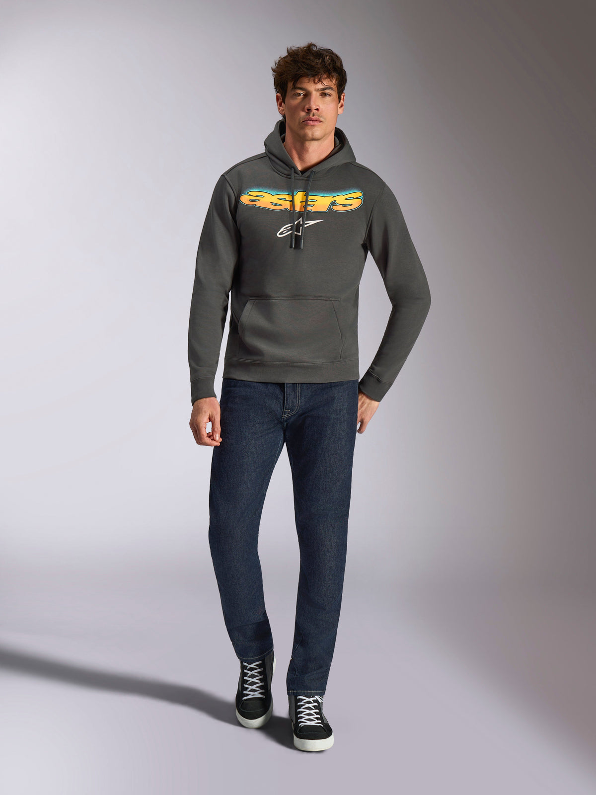 Alpinestars Ellipsoid Hoodie, abbigliamento sportivo casual, grigio gunmetal con logo Astars a gradiente arancione e giallo e simbolo bianco, cappuccio con cordino, indossata da un modello maschile con jeans blu scuro e sneakers nere, streetwear moderno ispirato al motorsport