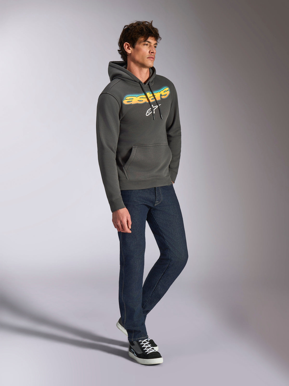 Felpa con cappuccio Alpinestars Ellipsoid, pullover in pile grigio canna di fucile, scritta frontale curva Astars con gradiente blu e giallo e logo bianco, abbinata a jeans scuri e sneakers nere, abbigliamento casual streetwear.