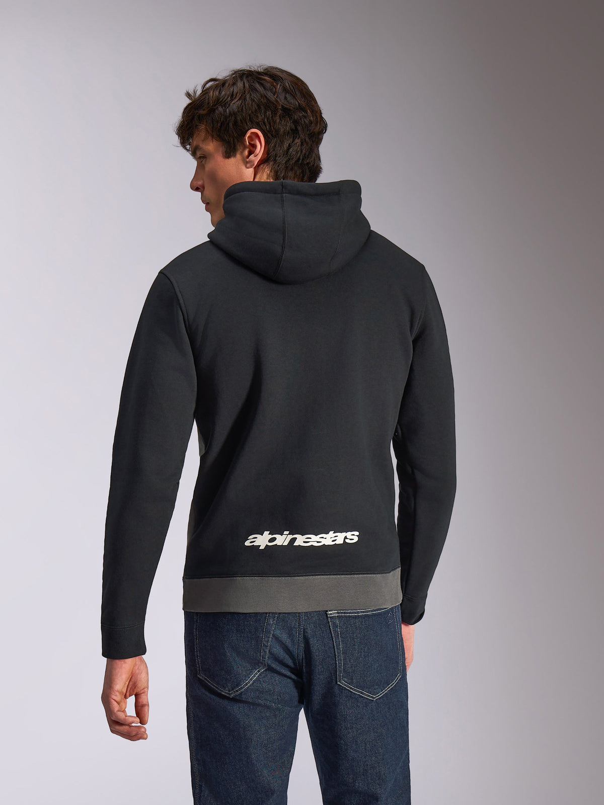 Alpinestars Parallel Hoodie, felpa casual sportswear, nero e gunmetal, vista posteriore con logo Alpinestars bianco sulla parte inferiore della schiena, design a blocchi di colore con bordo grigio, indossata con jeans in denim scuro