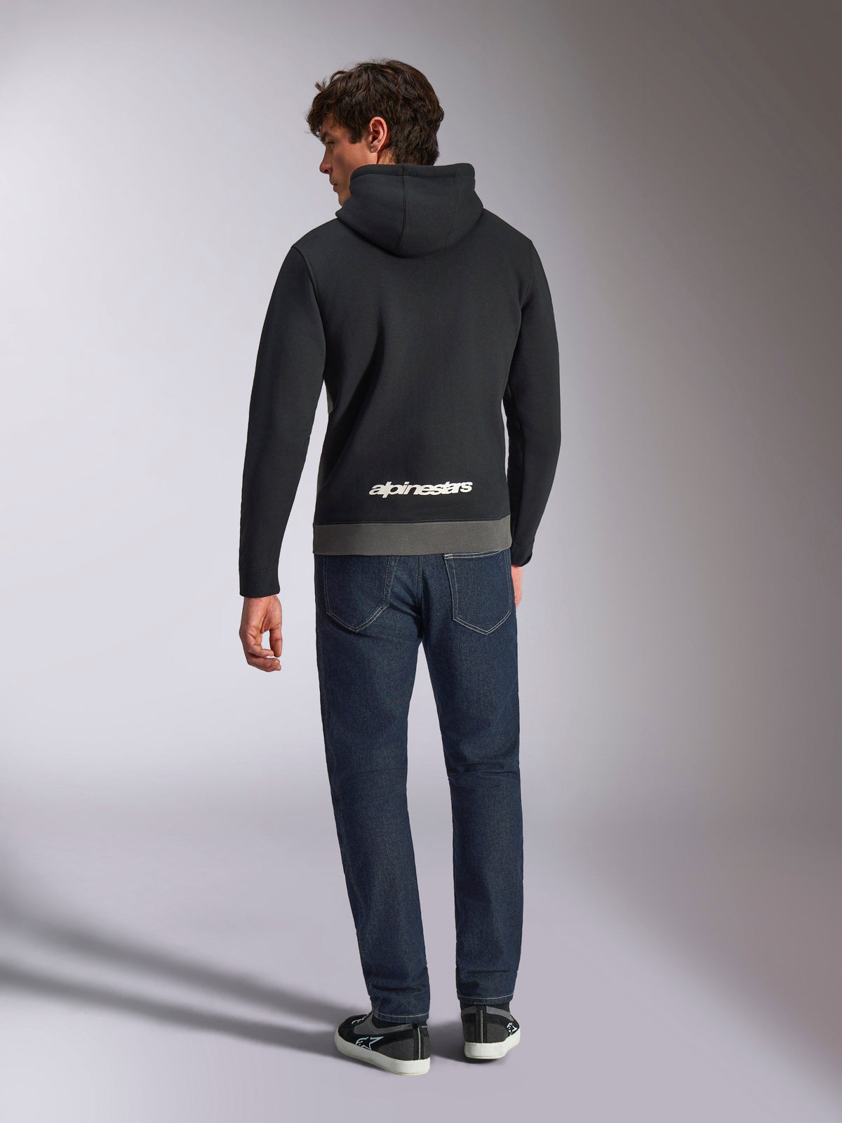 Felpa con cappuccio Alpinestars Parallel, vista posteriore di un uomo che indossa un pullover in pile a blocchi di colore nero e gunmetal, logo Alpinestars bianco sulla parte inferiore della schiena, abbinata a jeans blu scuro, streetwear casual ispirato al motorsport
