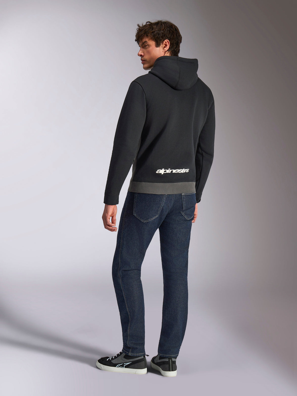 Felpa con cappuccio Alpinestars Parallel, vista posteriore del modello che indossa il pullover nero e grigio canna di fucile, con logo Alpinestars bianco sulla parte inferiore della schiena, abbinata a jeans blu scuro, streetwear casual d'ispirazione motorsport