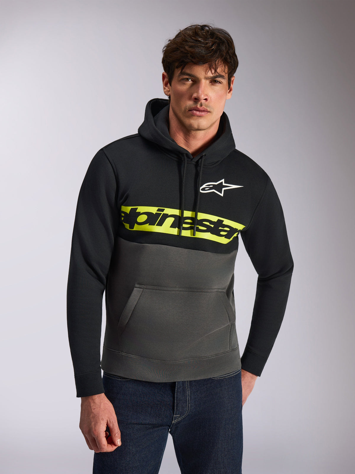 Alpinestars Parallel Hoodie, felpa con cappuccio casual, a blocchi di colore nero e grigio canna di fucile, striscia gialla brillante sul petto con logo Alpinestars, logo Astars bianco, indossata da un modello con jeans blu, design moderno ispirato al motorsport