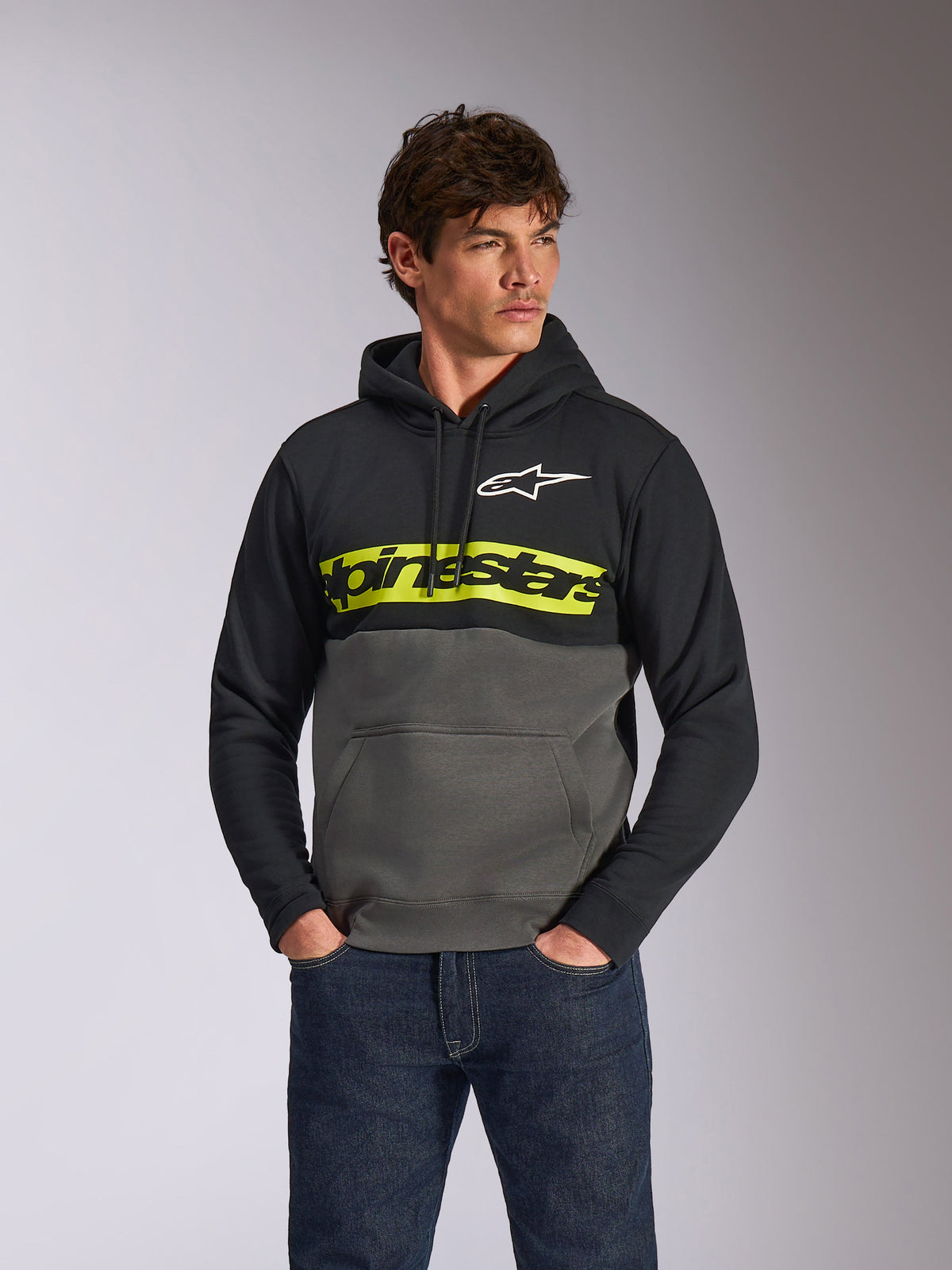 Alpinestars Parallel Hoodie, felpa pullover casual a blocchi di colore, nero e grigio canna di fucile con una barra gialla vivace sul petto, logo Alpinestars e logo a stella bianco, indossata da un modello con jeans blu scuro