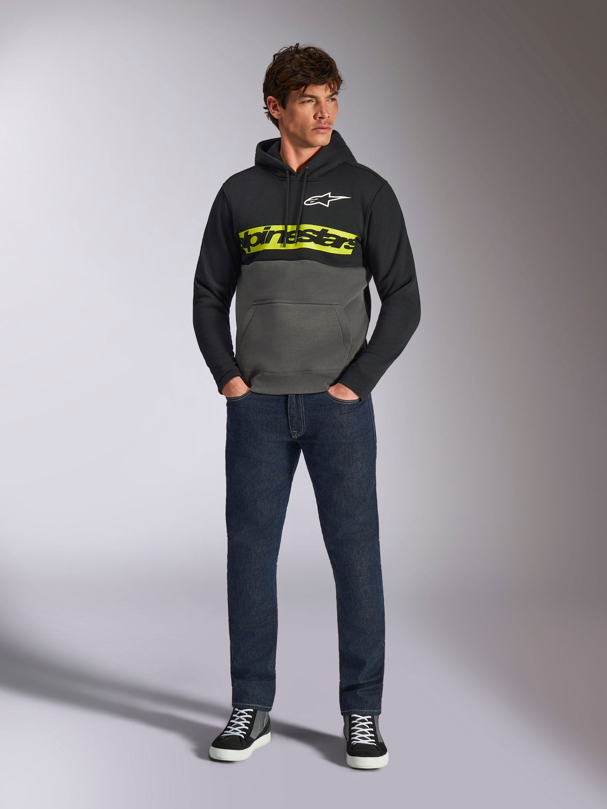 Alpinestars Parallel Hoodie, felpa con cappuccio a blocchi di colore, nero e grigio gunmetal, logo Alpinestars giallo acceso e logo a stella bianco sul petto, indossata da un modello con jeans blu scuro e sneakers nere, streetwear casual motorsport