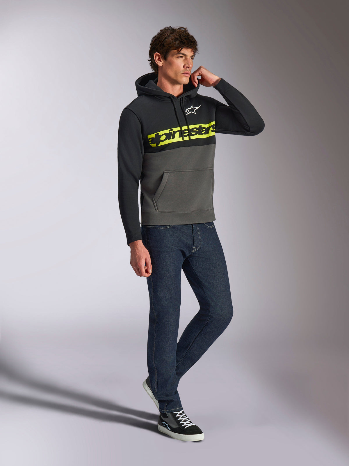 Felpa con cappuccio Alpinestars Parallel, streetwear casual, design a blocchi di colore nero e grigio gunmetal, logo word Alpinestars giallo neon sul petto, logo Astars bianco, cappuccio con cordino, indossata con jeans scuri e sneakers