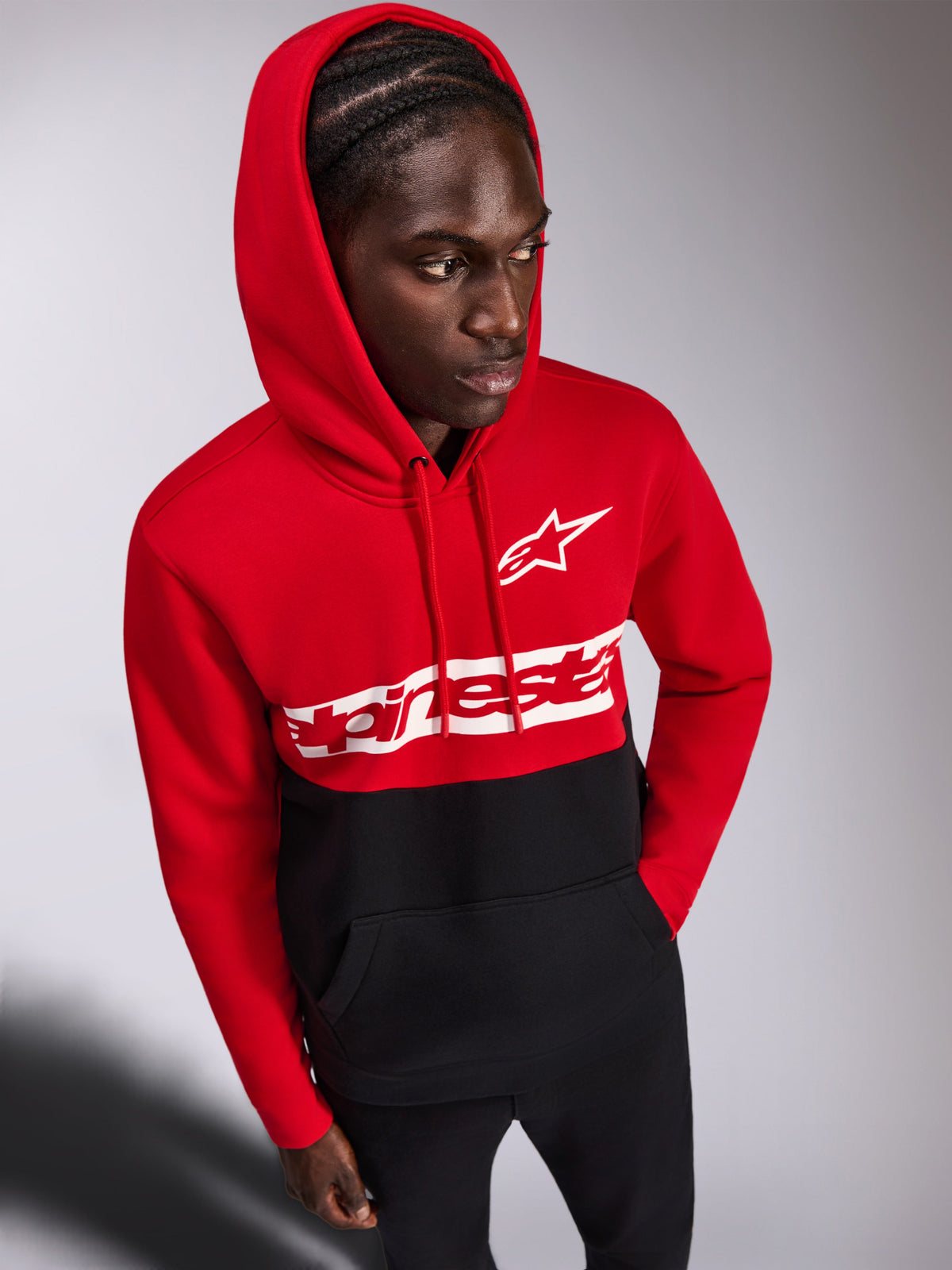 Felpa con cappuccio Alpinestars Parallel, felpa casual pullover, design a blocchi di colore rosso e nero, logo Alpinestars Word e logo Astars bianchi sul petto, indossata da un uomo con il cappuccio sollevato e pantaloni neri, streetwear ispirato al motorsport