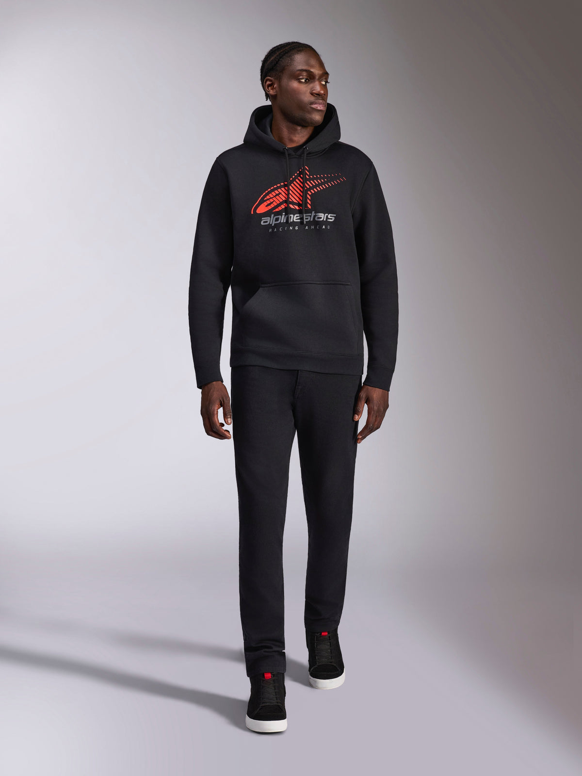 Alpinestars Pulse Hoodie, felpa con cappuccio casual streetwear nera, logo Astars combo rosso e grigio con linea di velocità sfumata sul petto, cappuccio con cordino, tasca a marsupio anteriore, maniche lunghe, abbinata a pantaloni neri e sneakers, design ispirato al motorsport