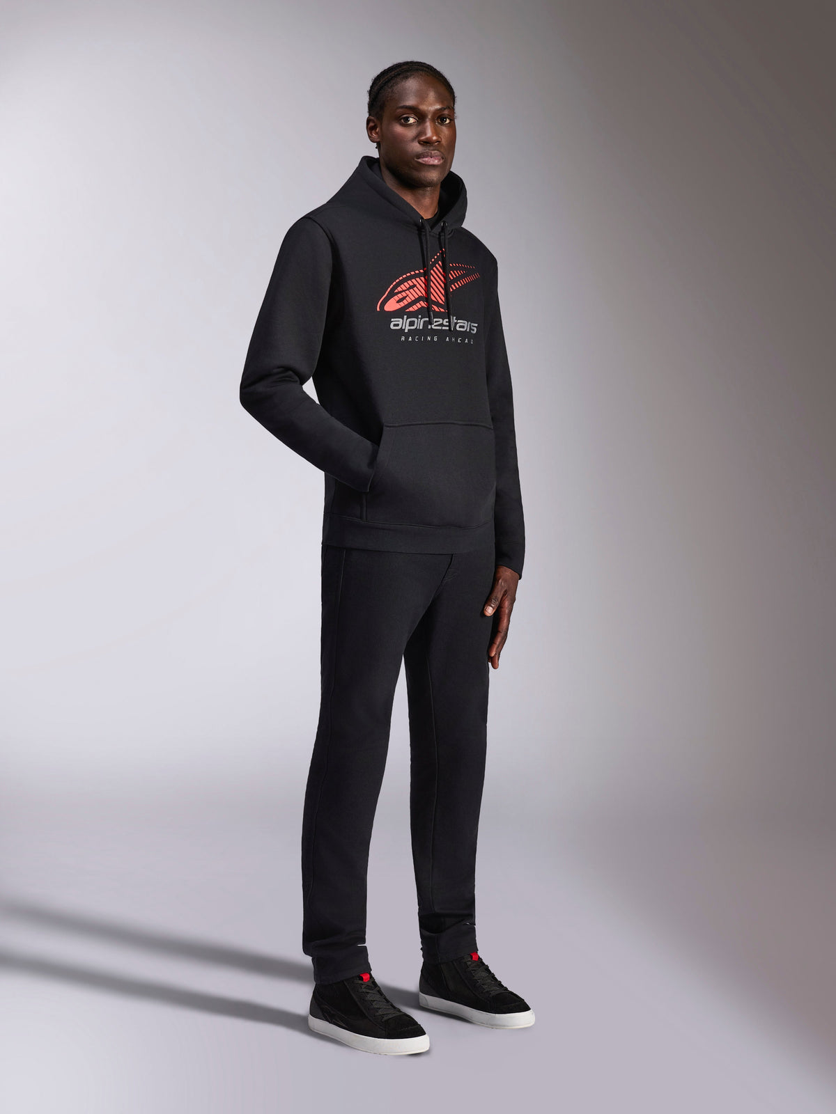 Alpinestars Pulse Hoodie, felpa nera con cappuccio, con logo Astars rosso e bianco, linea di velocità sfumata e scritta Racing Ahead, indossata con pantaloni e sneaker neri coordinati, streetwear casual ispirato al motorsport.