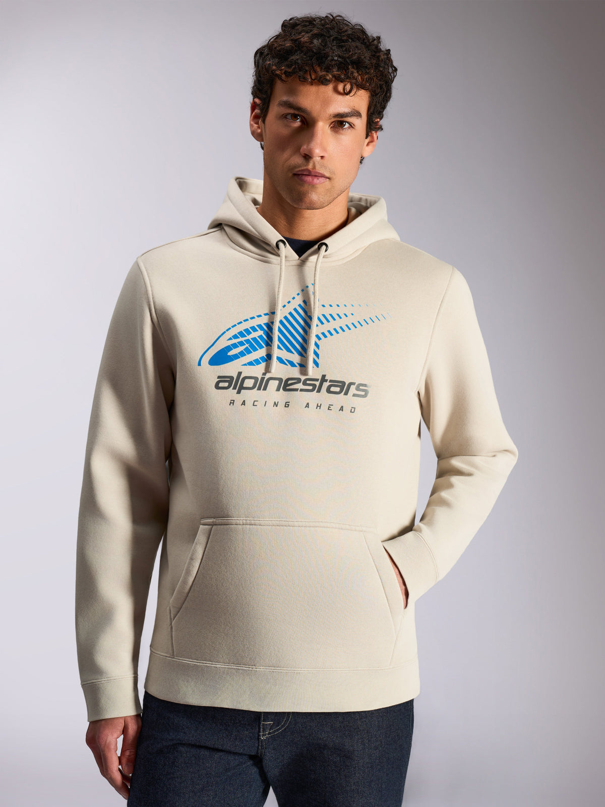 Felpa con cappuccio Alpinestars Pulse, streetwear casual, colore sabbia, logo frontale Astars Combo blu e grigio con sfumatura a linee di velocità e scritta 'Racing Ahead', cappuccio con cordino, tasca a marsupio, indossata da un modello con jeans scuri