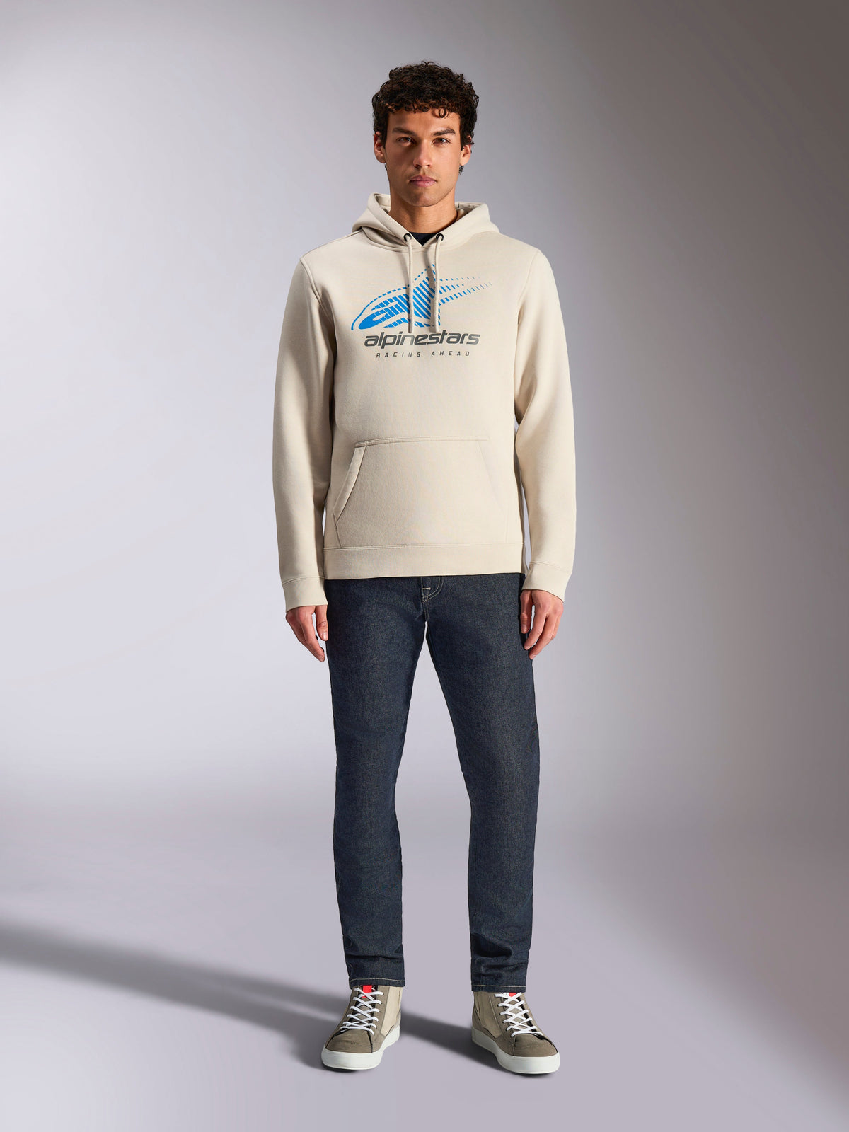 Felpa con cappuccio Alpinestars Pulse, bianco sporco, pullover a maniche lunghe con cappuccio e cordino, logo Astars Combo blu e nero con linea di velocità sfumata, abbinata a jeans scuri e sneakers, streetwear casual motorsport