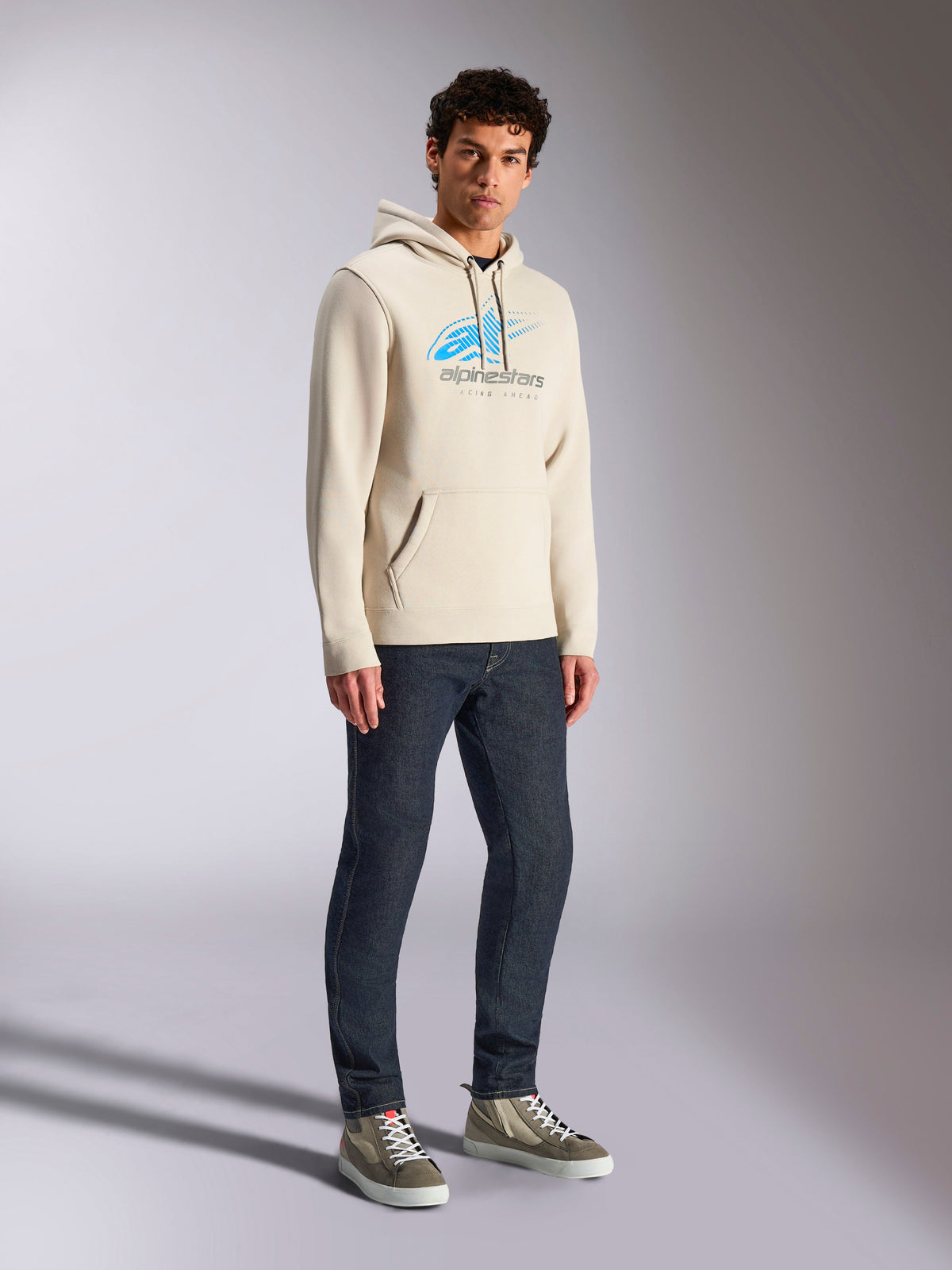 Alpinestars Felpa con cappuccio Pulse, streetwear casual Off White, felpa pullover con cappuccio con logo Astars blu e grigio e sfumatura a linee di velocità, tasca a marsupio, indossata da un modello con jeans in denim scuro e sneakers alte.